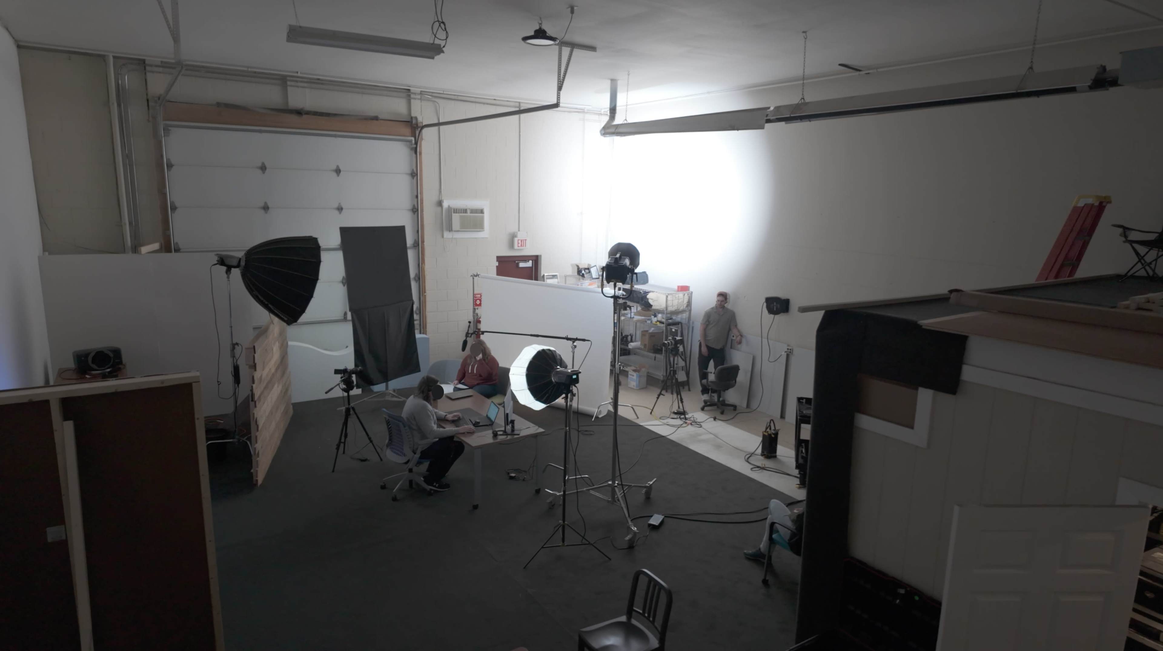 The image shows a photography studio with various lighting equipment set up and a person sitting at a table.
