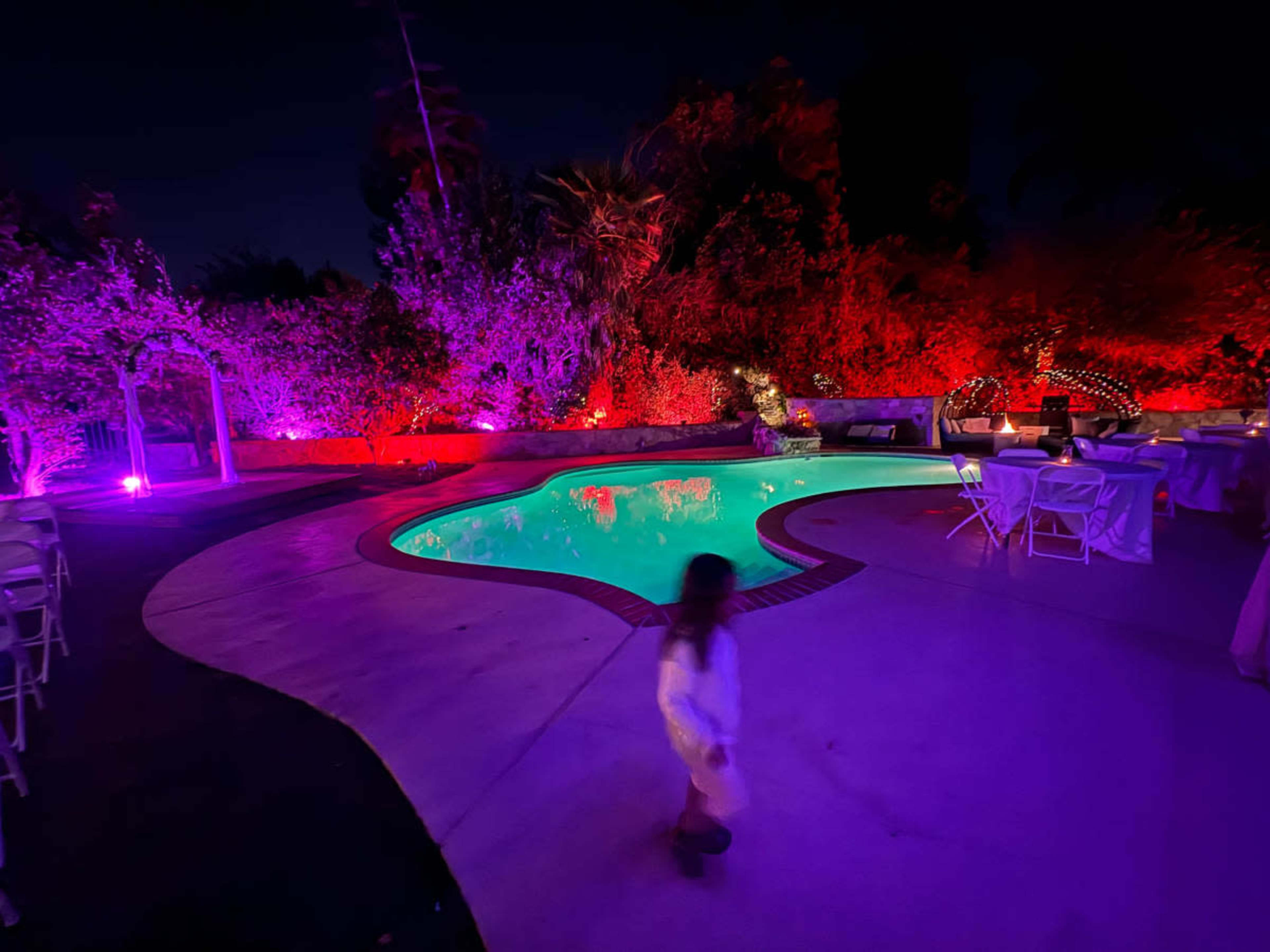 A brightly lit pool area features colorful lighting and empty seating, with a child walking near the edge of the water.