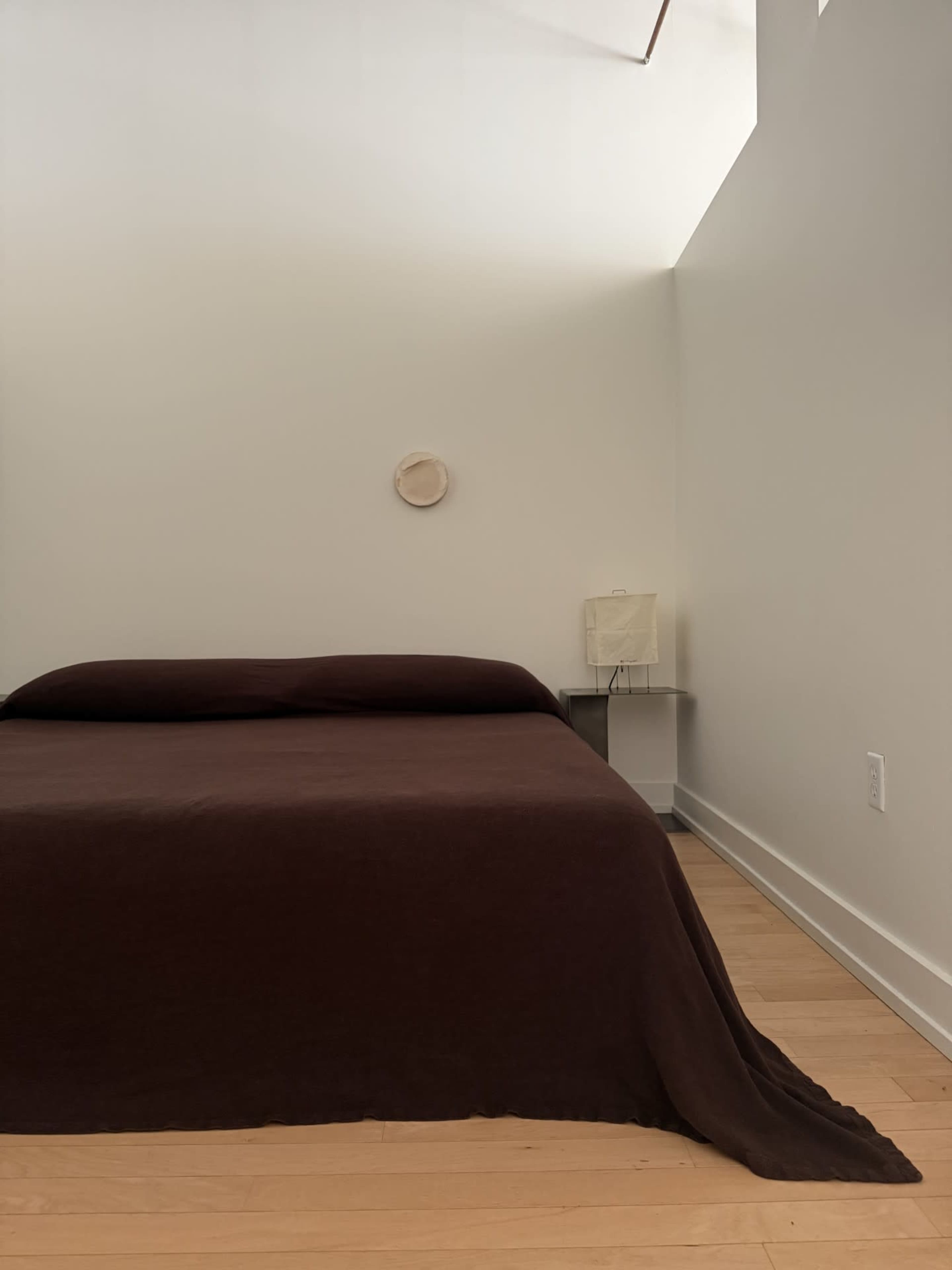 A minimalist bedroom features a brown bedspread on a bed with a small bedside table and a lamp in a softly lit space.
