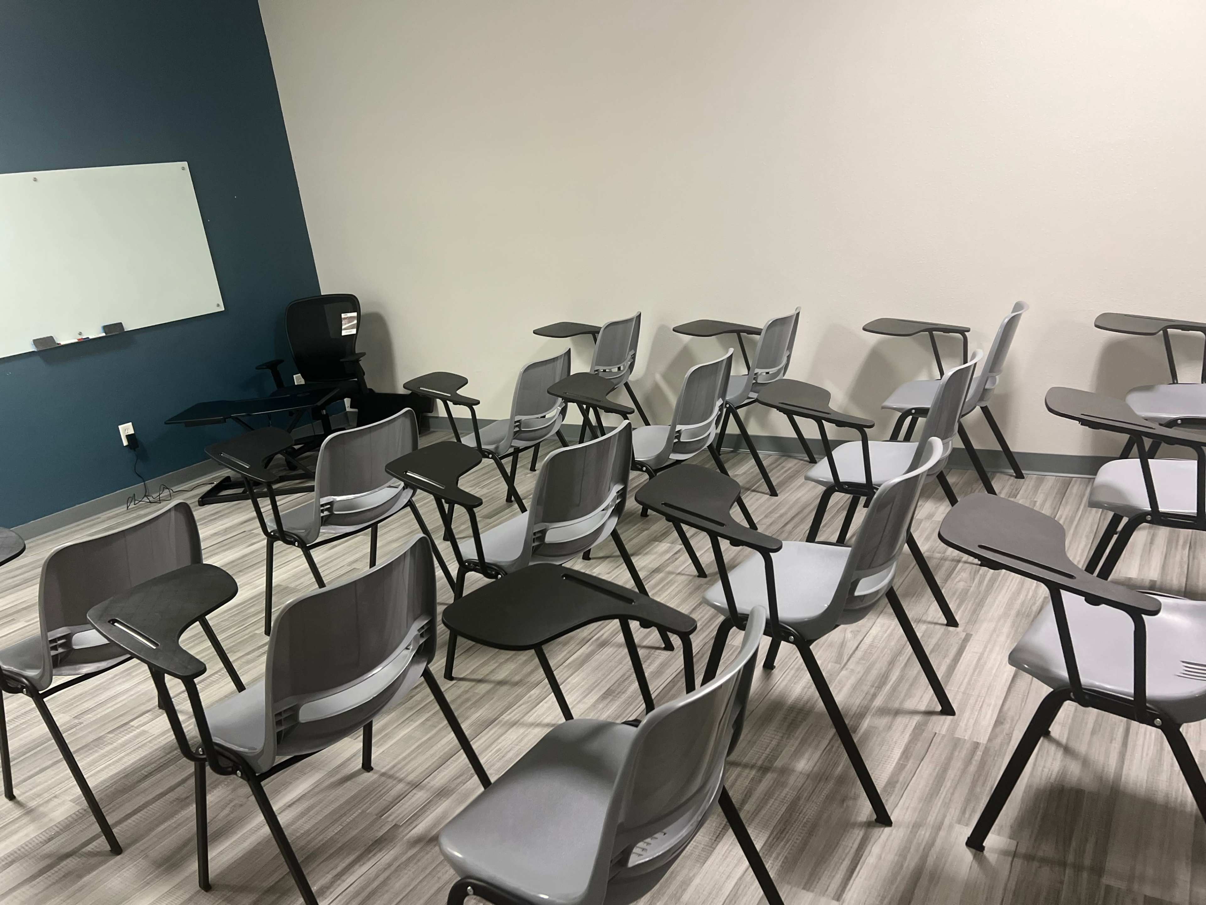 The image shows a classroom setting with several gray chairs arranged in a scattered formation around a central area, accompanied by a black office chair and a whiteboard on the wall.
