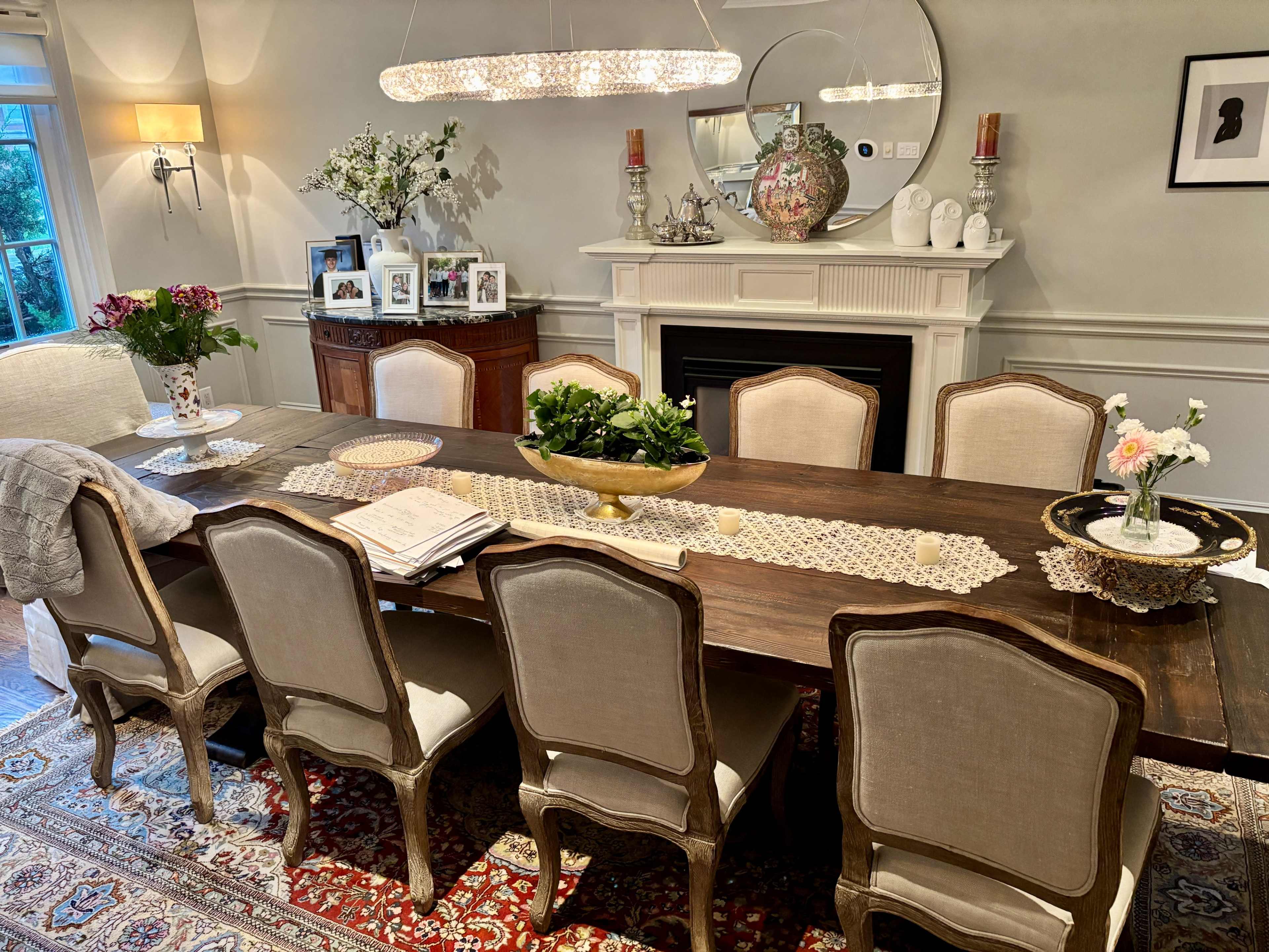 The image showcases a dining room featuring a long wooden table surrounded by upholstered chairs, with decorative items, flowers, and framed photographs on display.