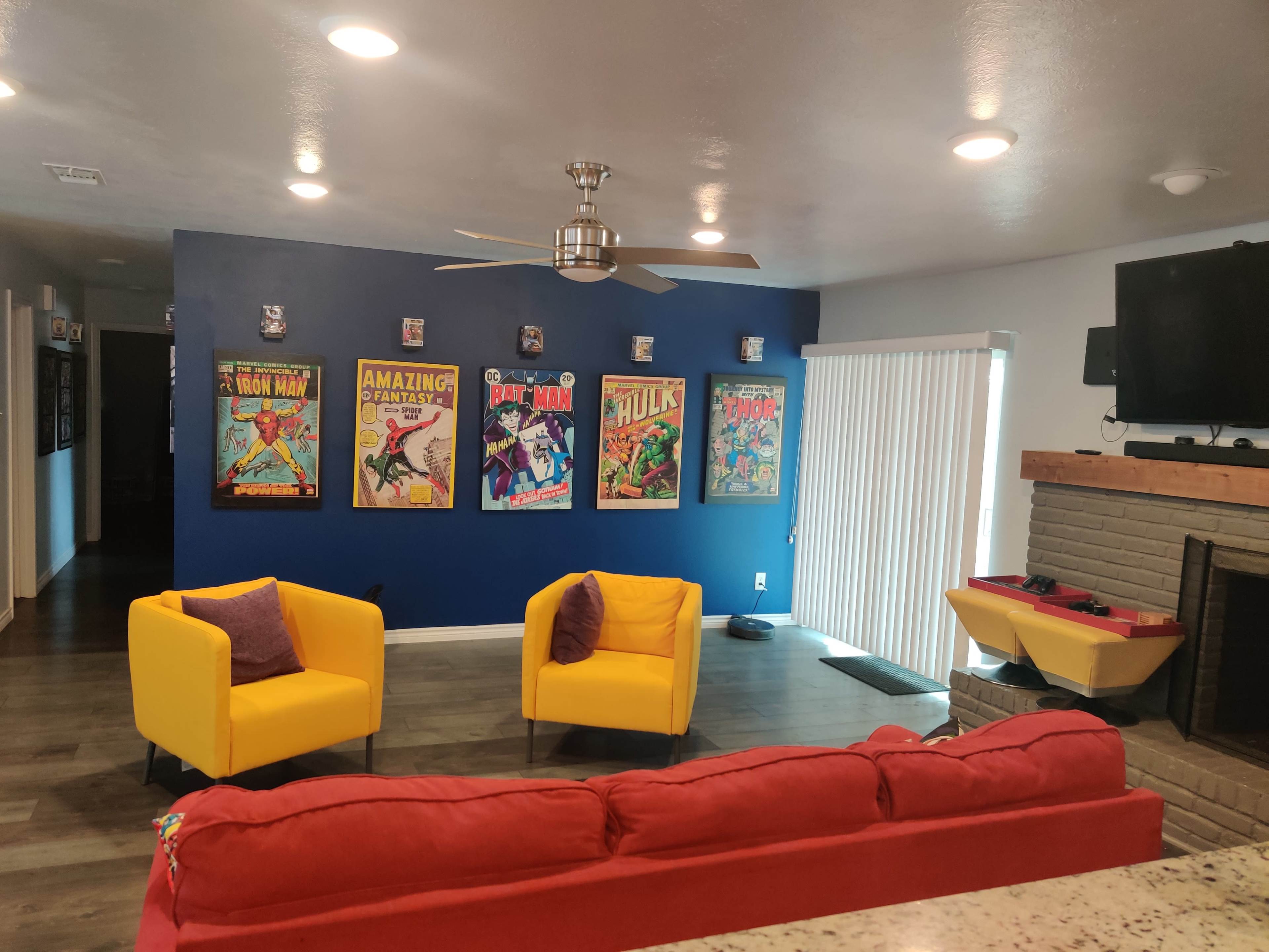 The room features a red sofa, two yellow armchairs, and a wall adorned with comic book posters, along with a fireplace and a television mounted above it.