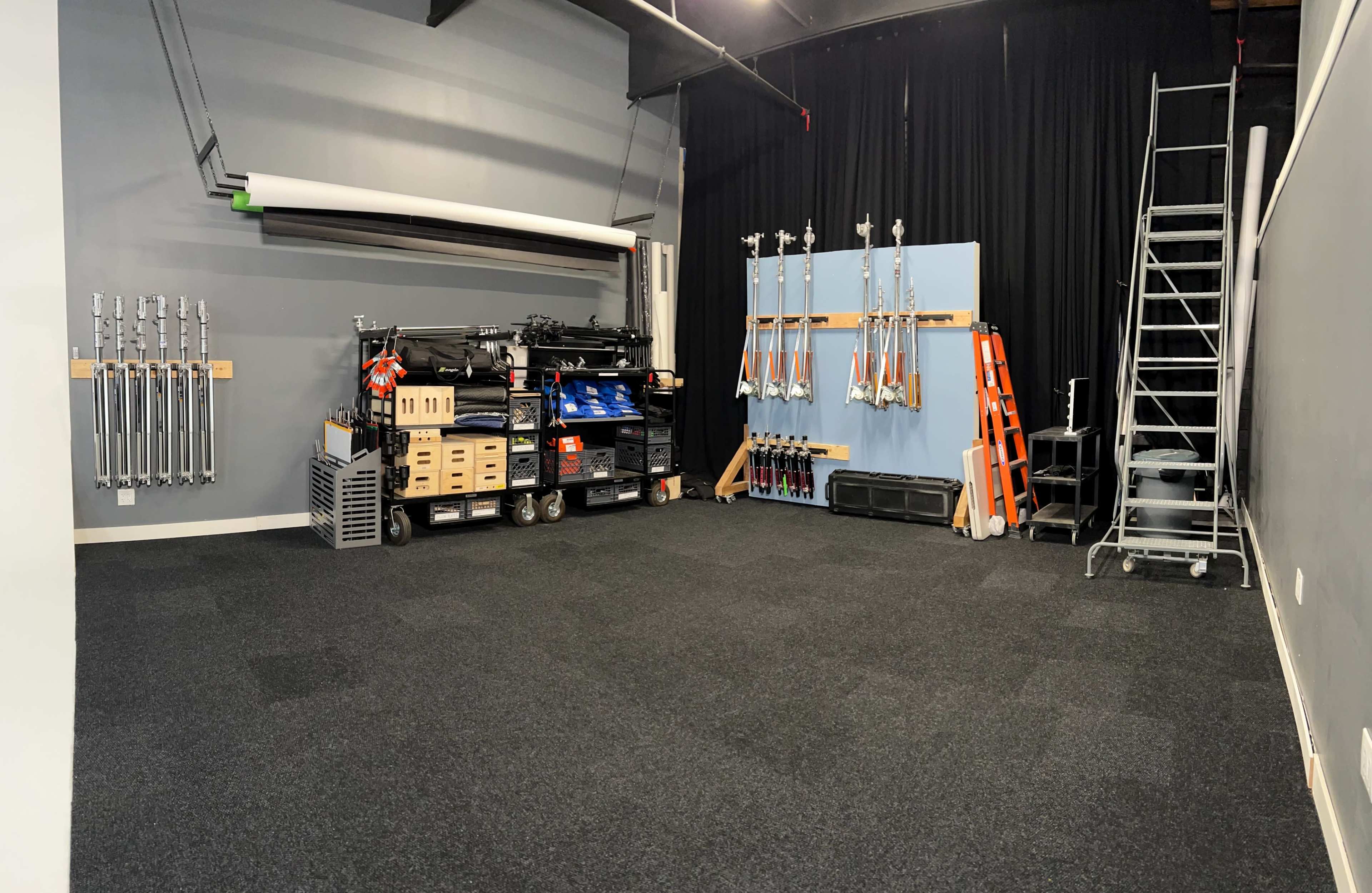 The image shows a spacious room with grey walls, black carpeting, and various equipment organized on shelves and carts, including lighting stands and a ladder.