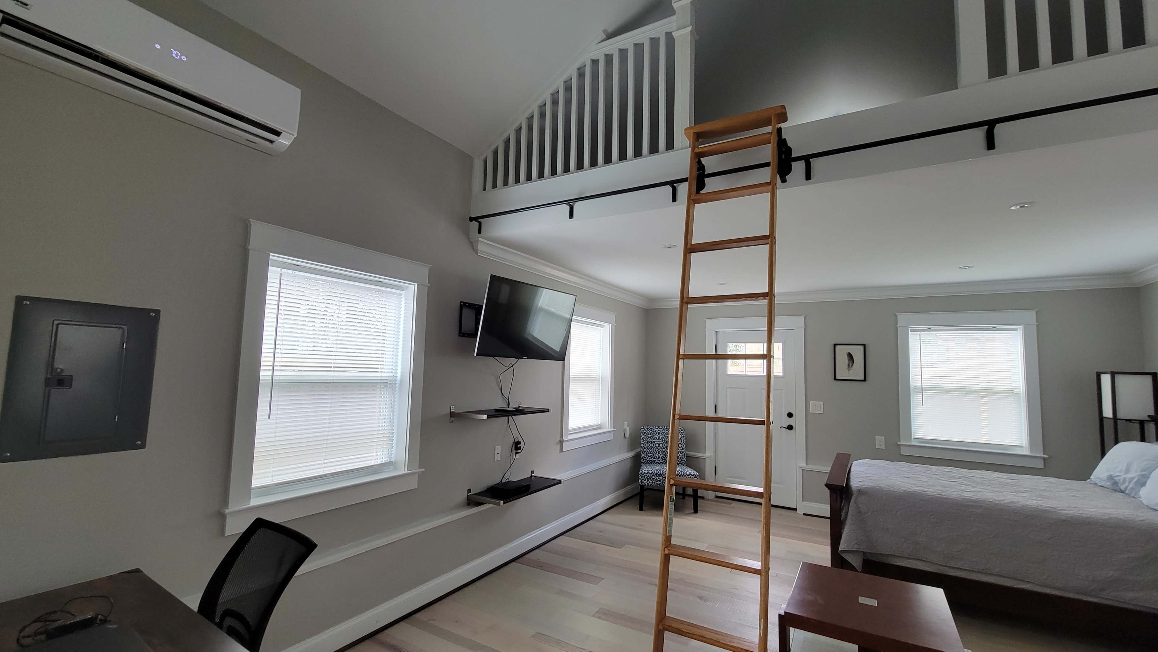 The image shows a modern room with a loft, featuring a ladder, a wall-mounted TV, a desk, and large windows allowing natural light.
