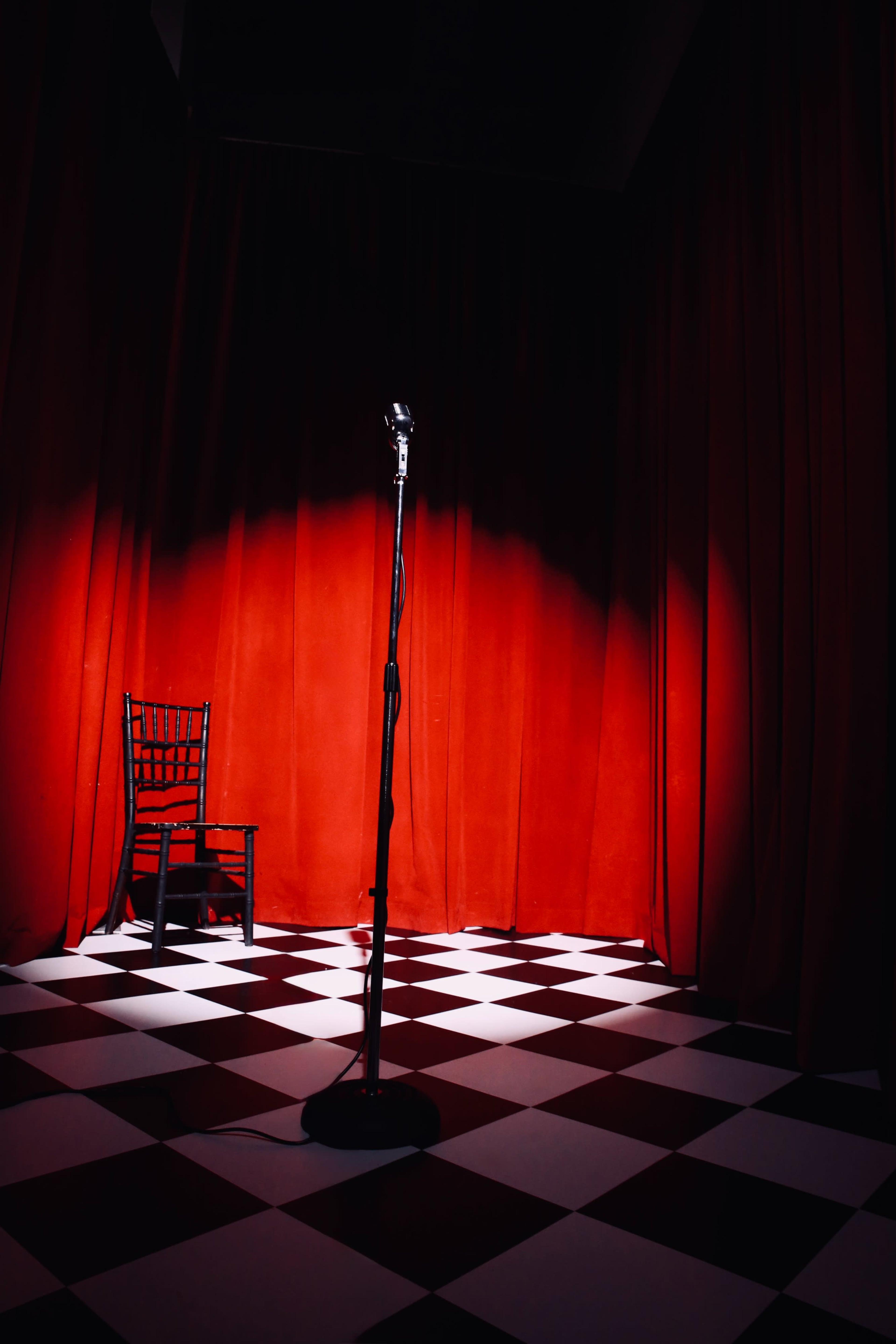 A microphone stand is positioned in front of a red curtain, with a single chair nearby on a checkered floor.
