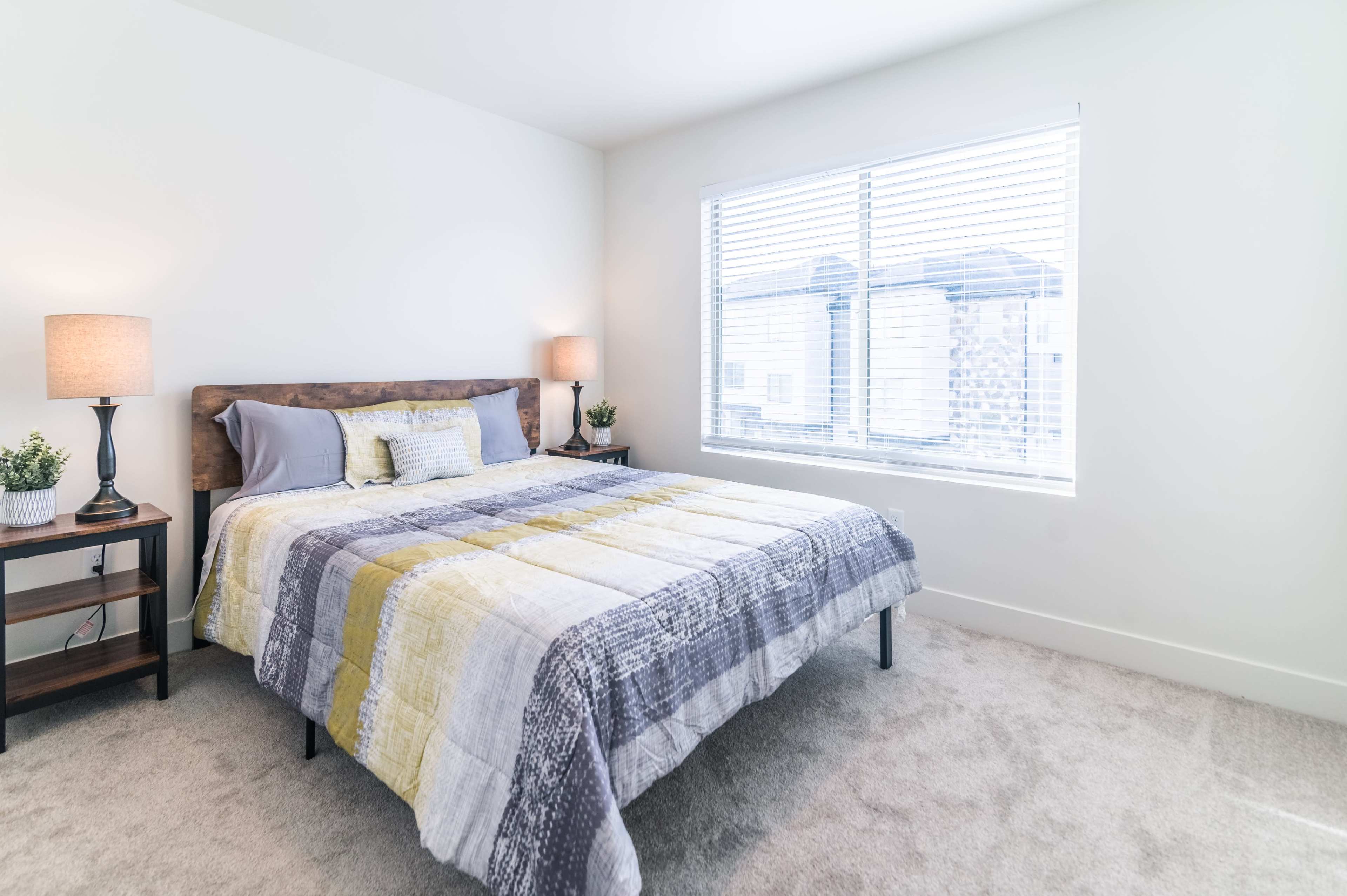 A well-lit bedroom featuring a bed with a patterned quilt, a nightstand with a lamp, and a window with blinds.