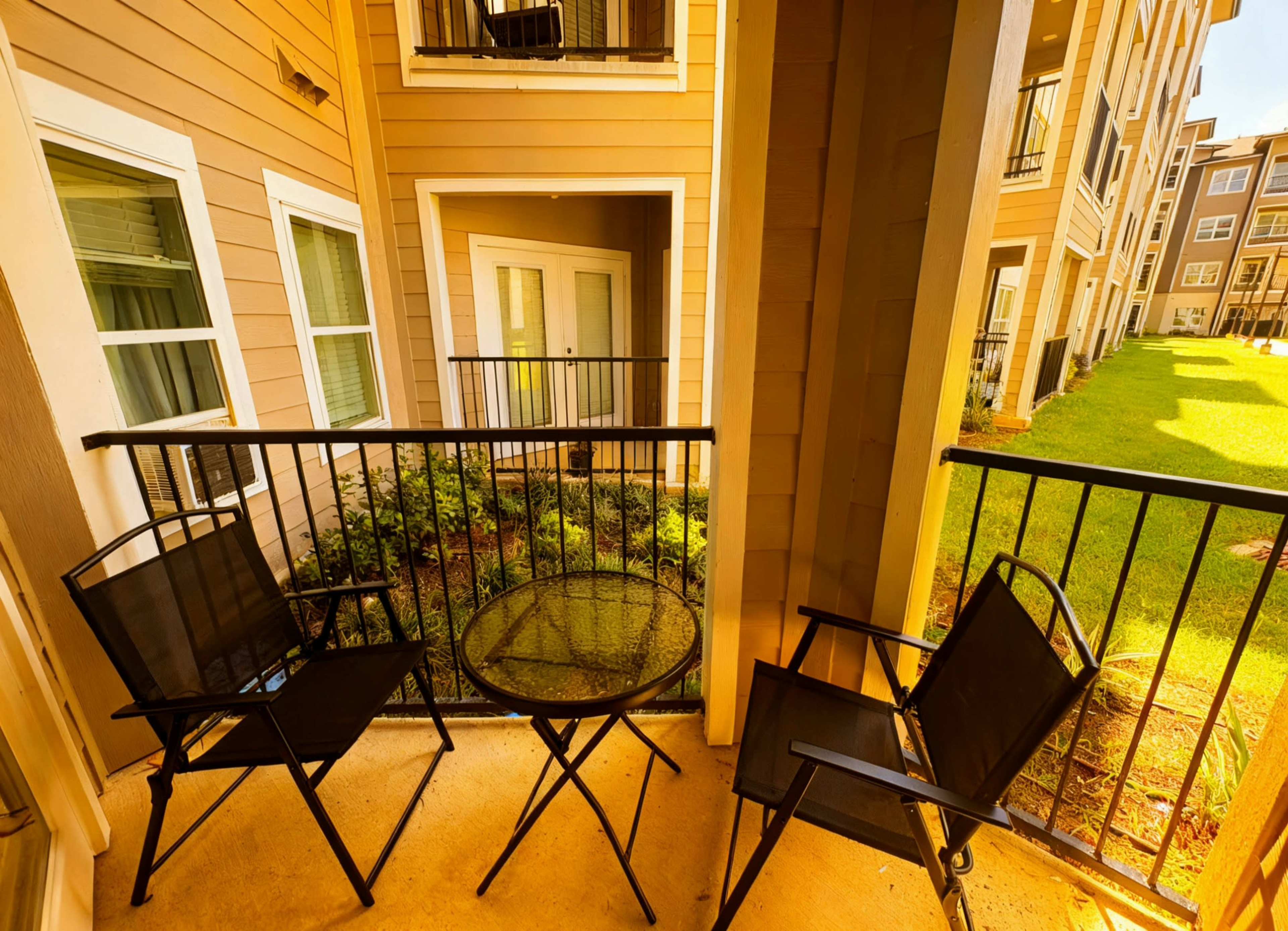 The image shows a small balcony with two black chairs and a round glass table, overlooking a grassy area between buildings.
