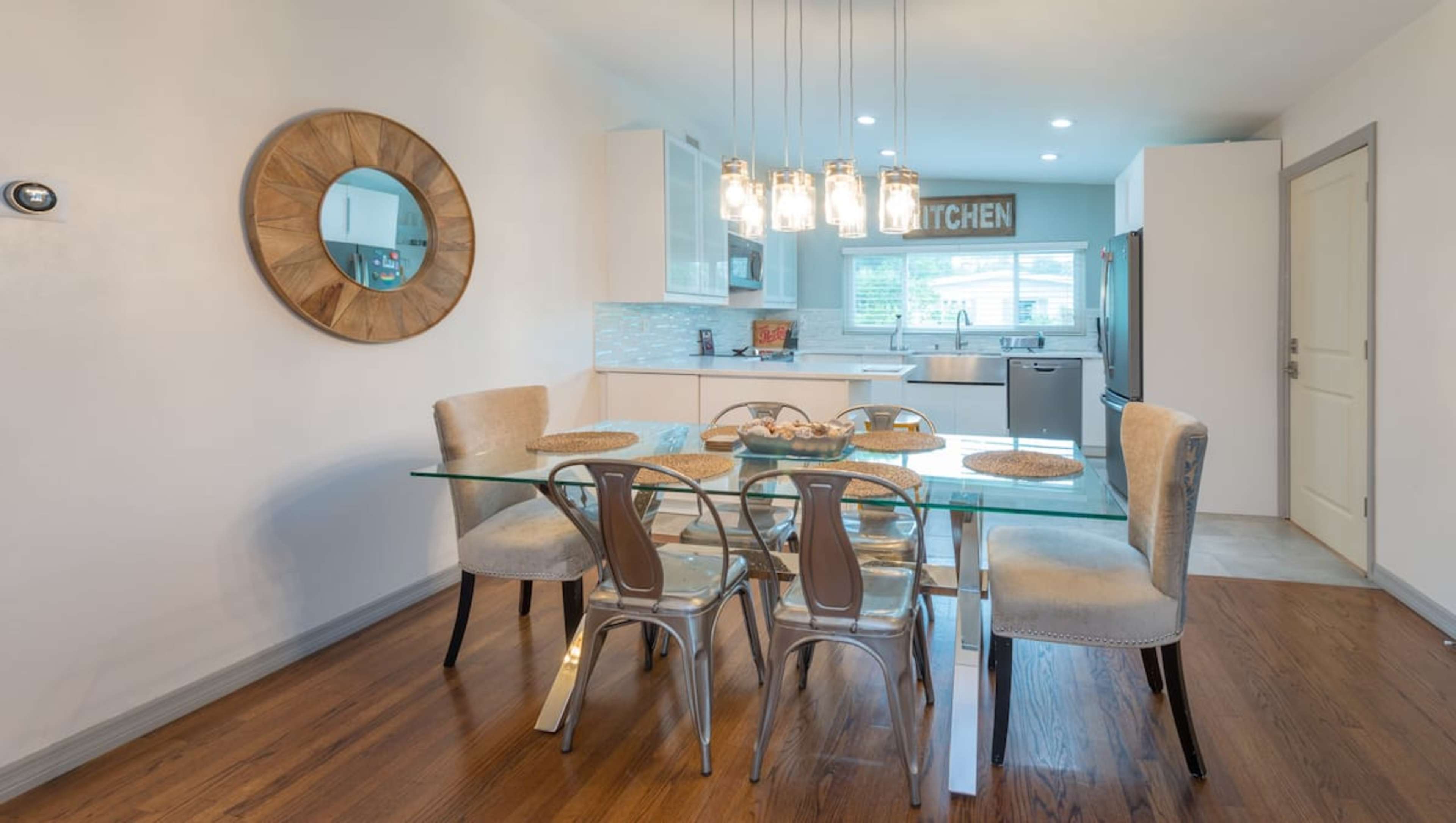 A modern dining area features a glass table with six chairs, a round mirror on the wall, and a kitchen visible in the background.