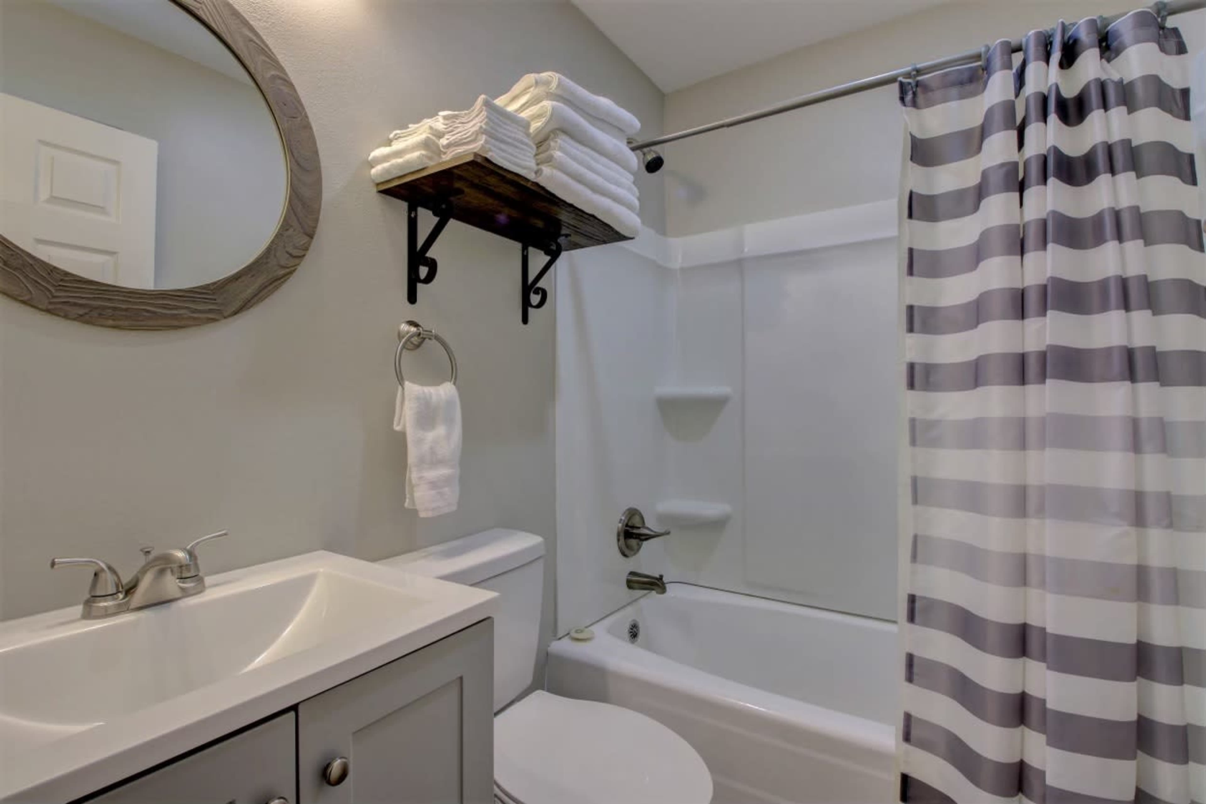 A bathroom with a shower curtain, a mirror, a sink with a cabinet, and neatly stacked towels on a shelf.