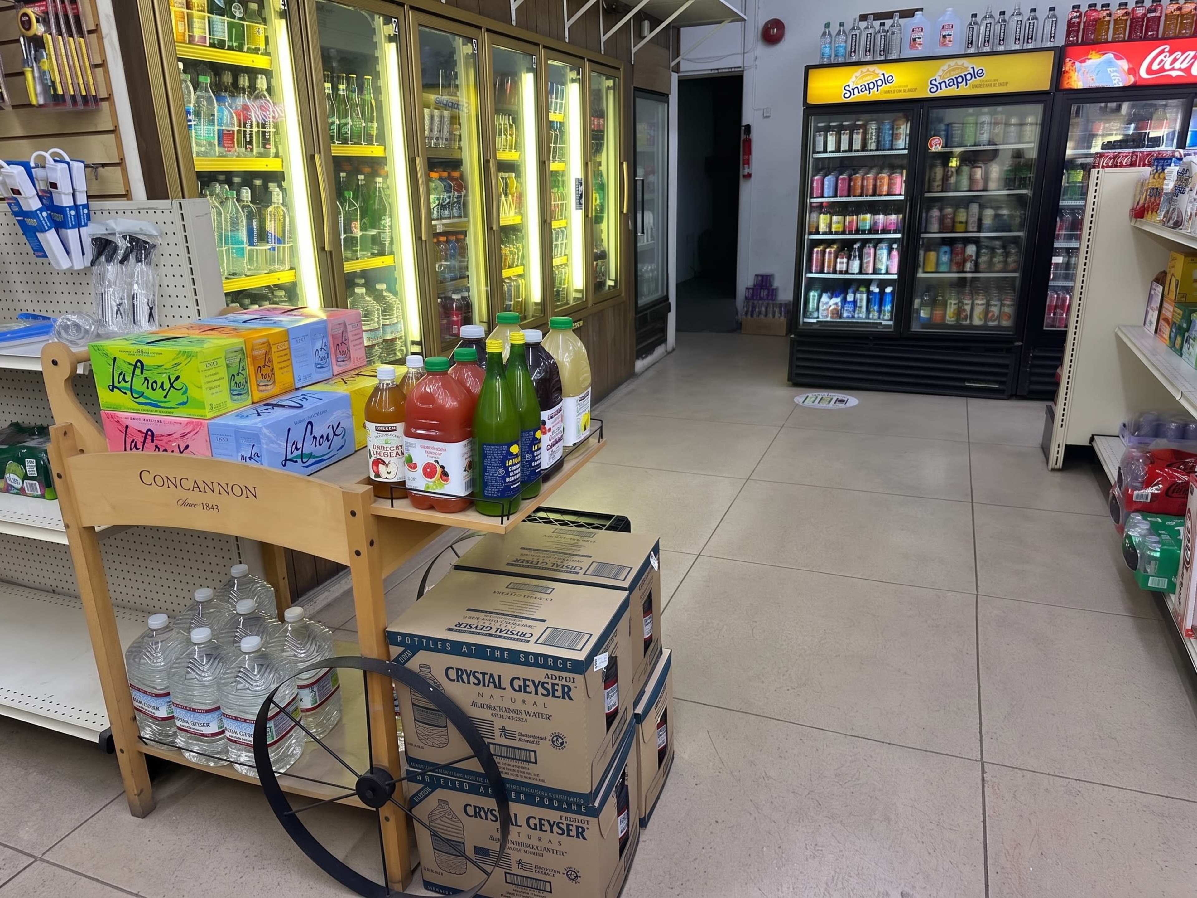 The image shows an interior of a convenience store with refrigerated beverage displays on one side and a display cart featuring juice and soda bottles in the foreground.
