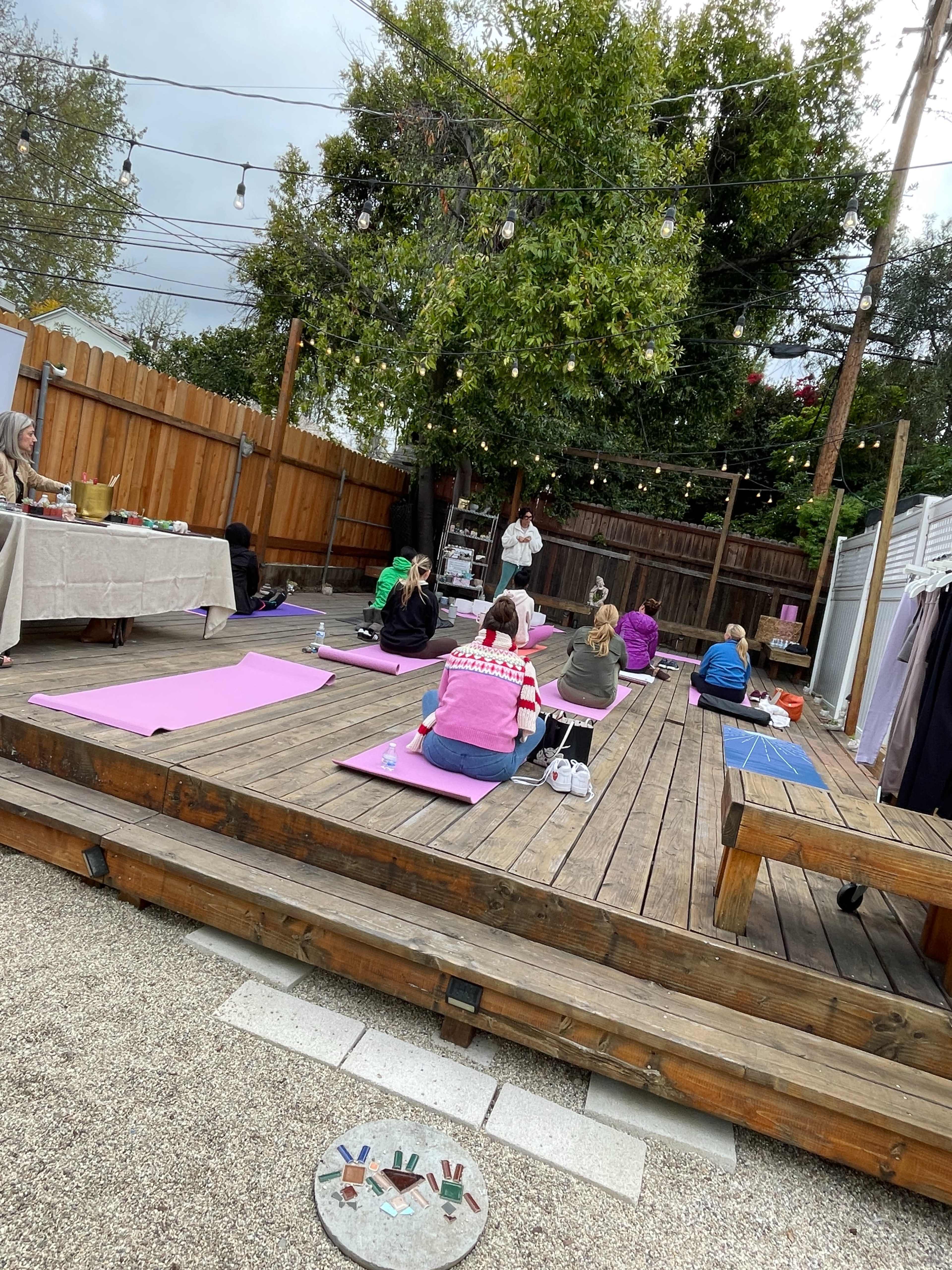 A group of people is participating in a yoga class in an outdoor wooden space with mats laid out on a deck beneath overhead string lights.