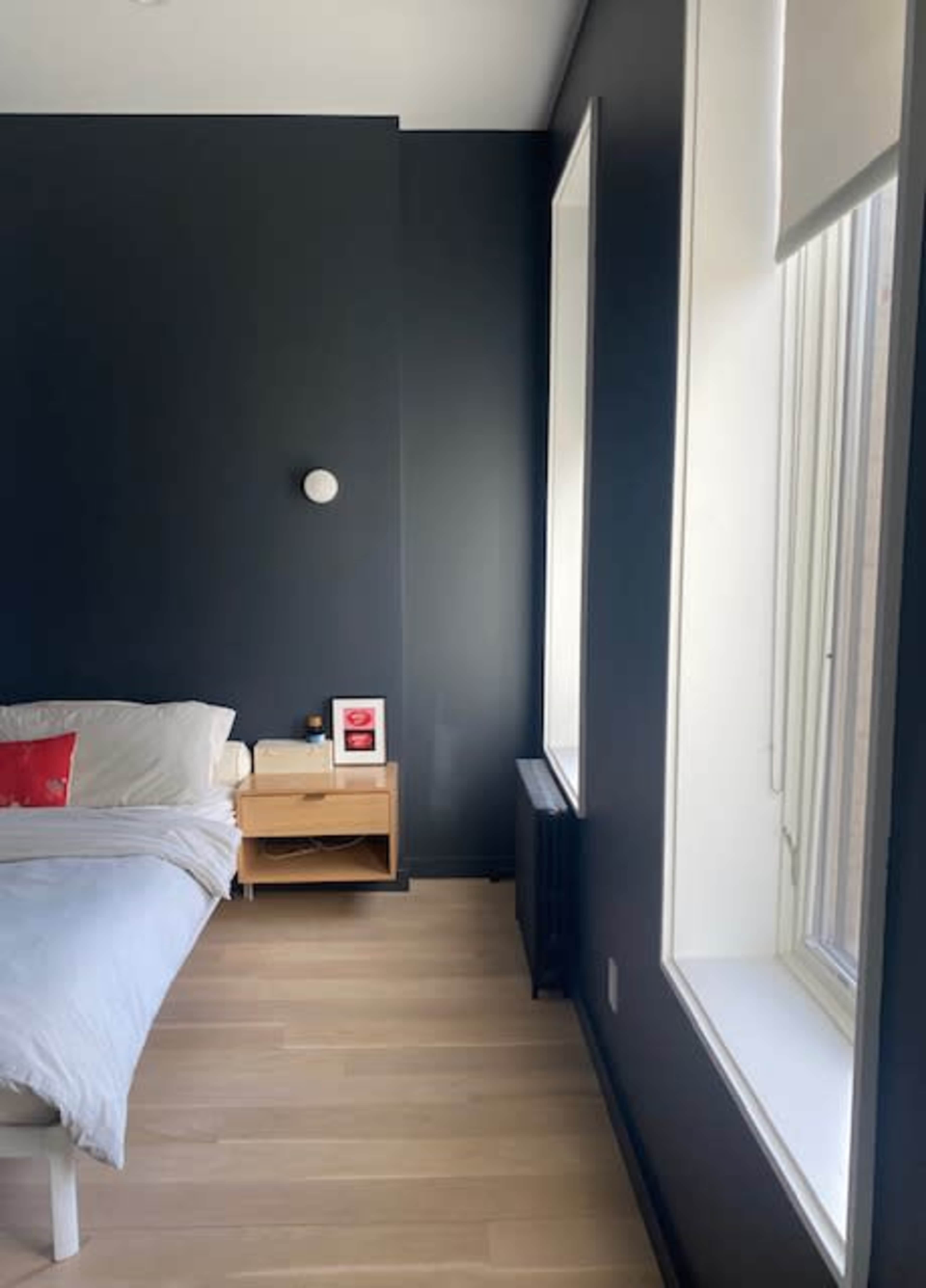 A modern bedroom with a bed on the left, a wooden nightstand beside it, and a large window on the right.