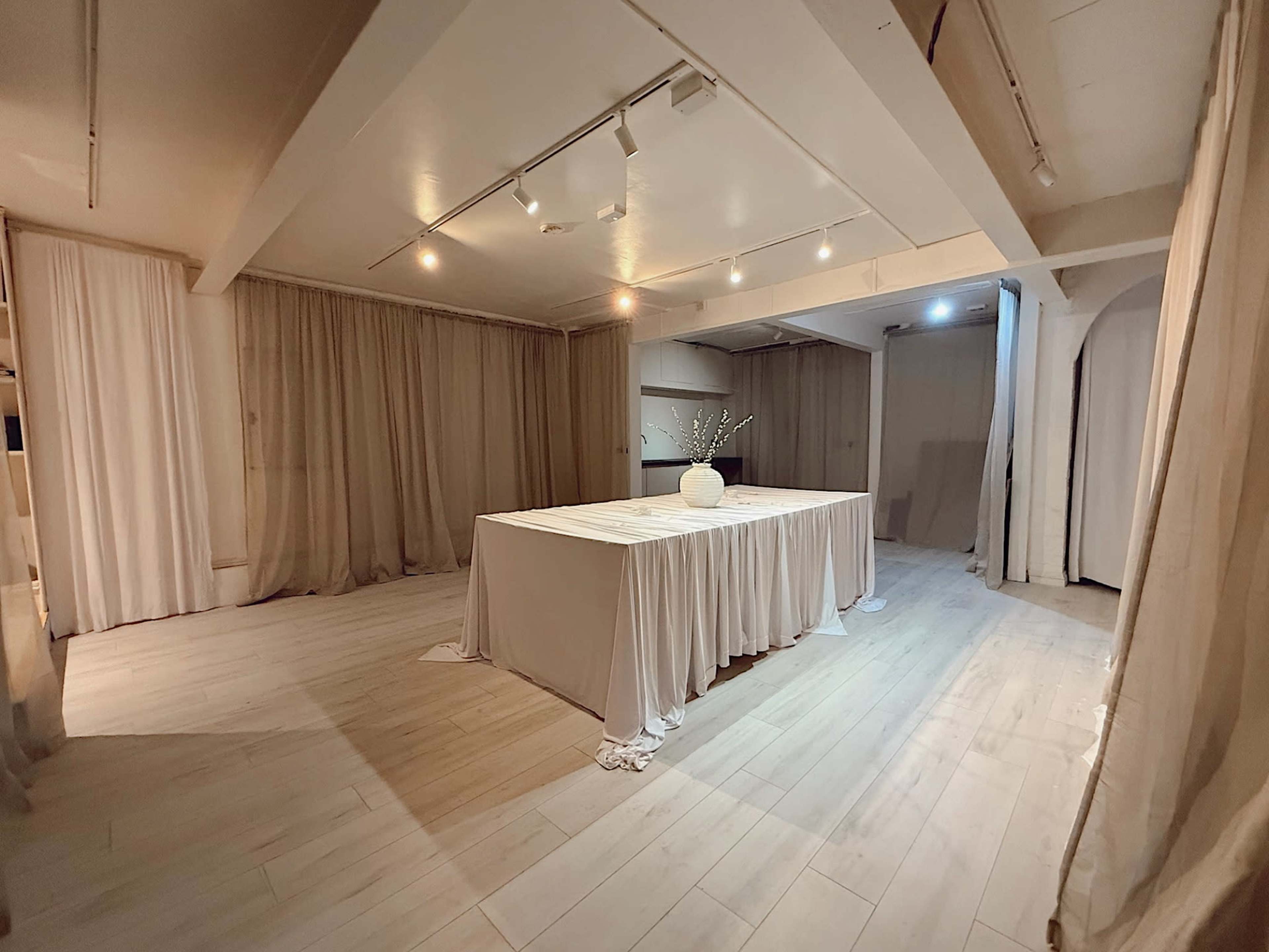 The image shows a spacious room with light-colored wooden flooring and soft, draped curtains, centered around a large, covered table.