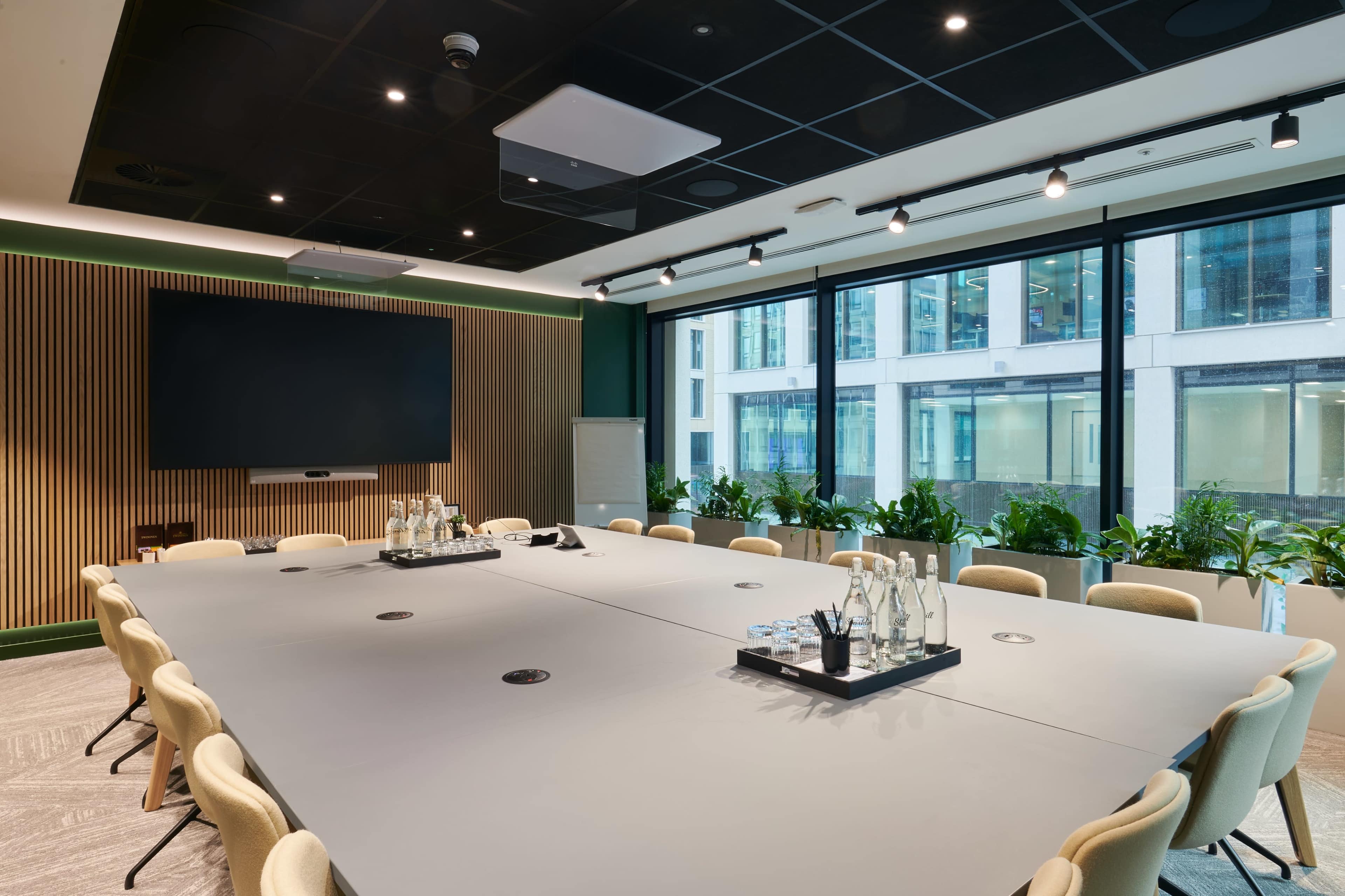 A modern conference room features a large table surrounded by chairs, with a screen on the wall and windows overlooking a cityscape.
