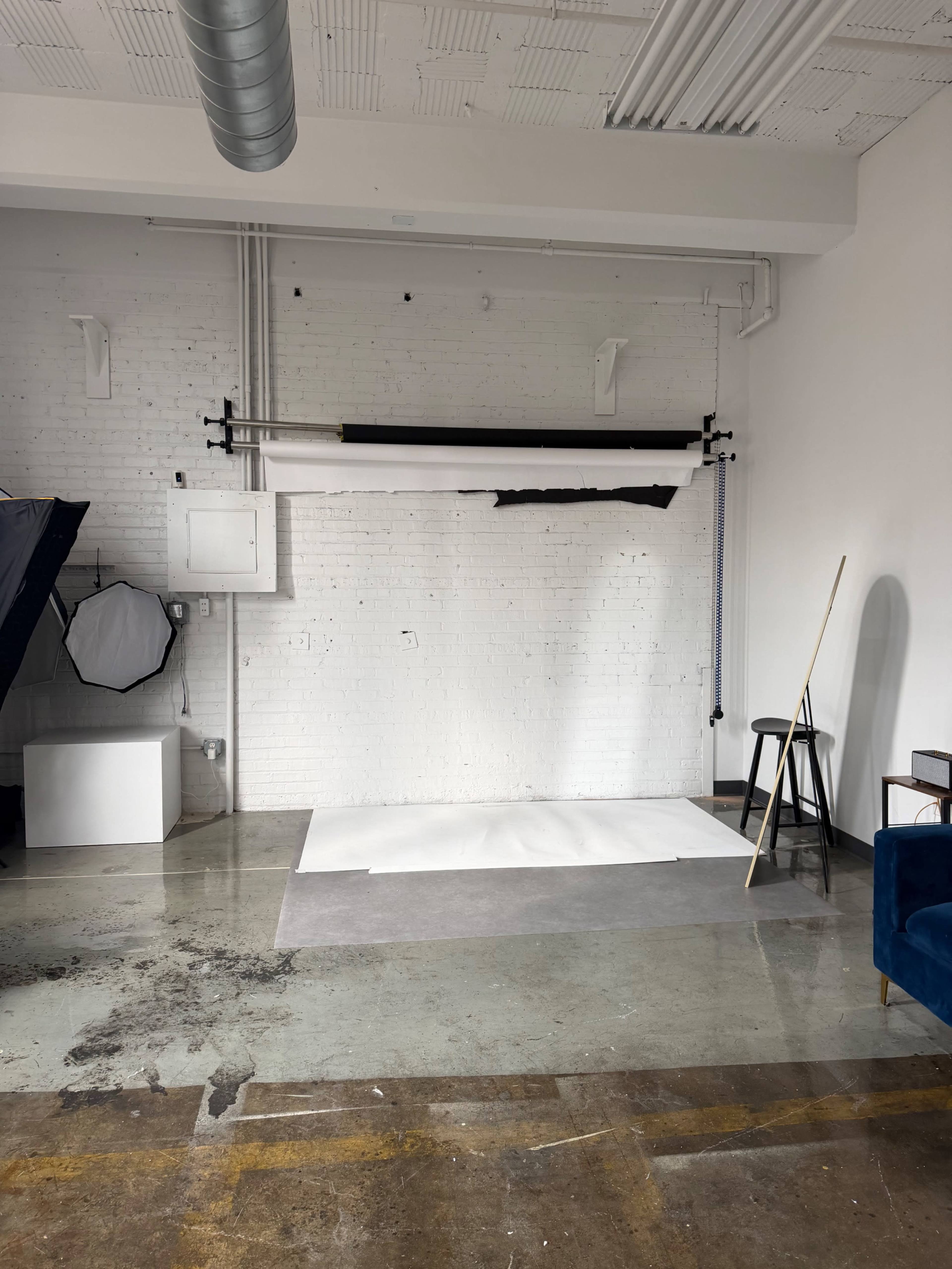 The image shows a photography studio with a clean, white backdrop on the wall, a roll of paper on a support system, and various studio equipment in the corner.