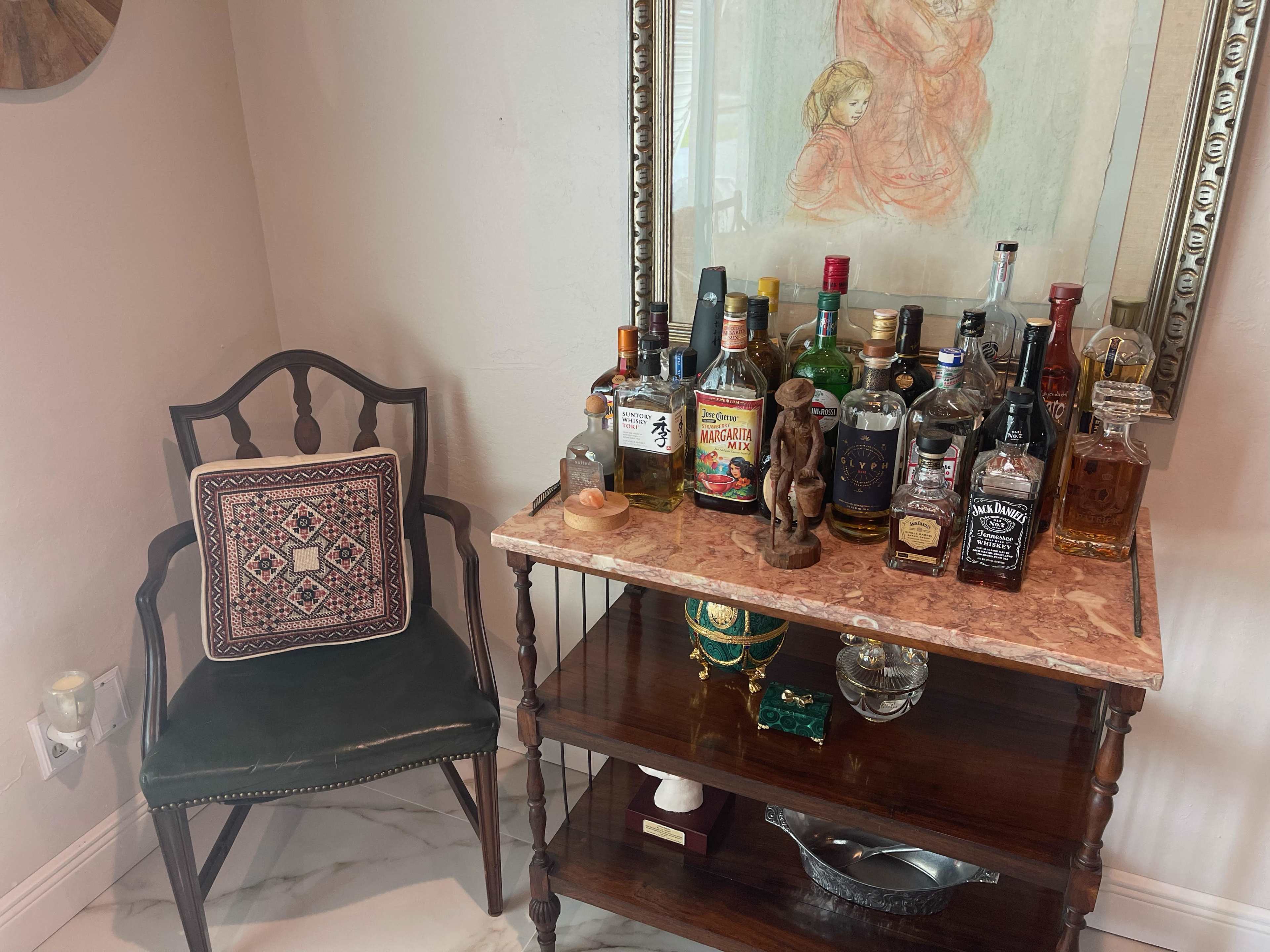 A wooden table topped with various bottles of liquor is positioned beside a chair and a framed artwork featuring two figures.