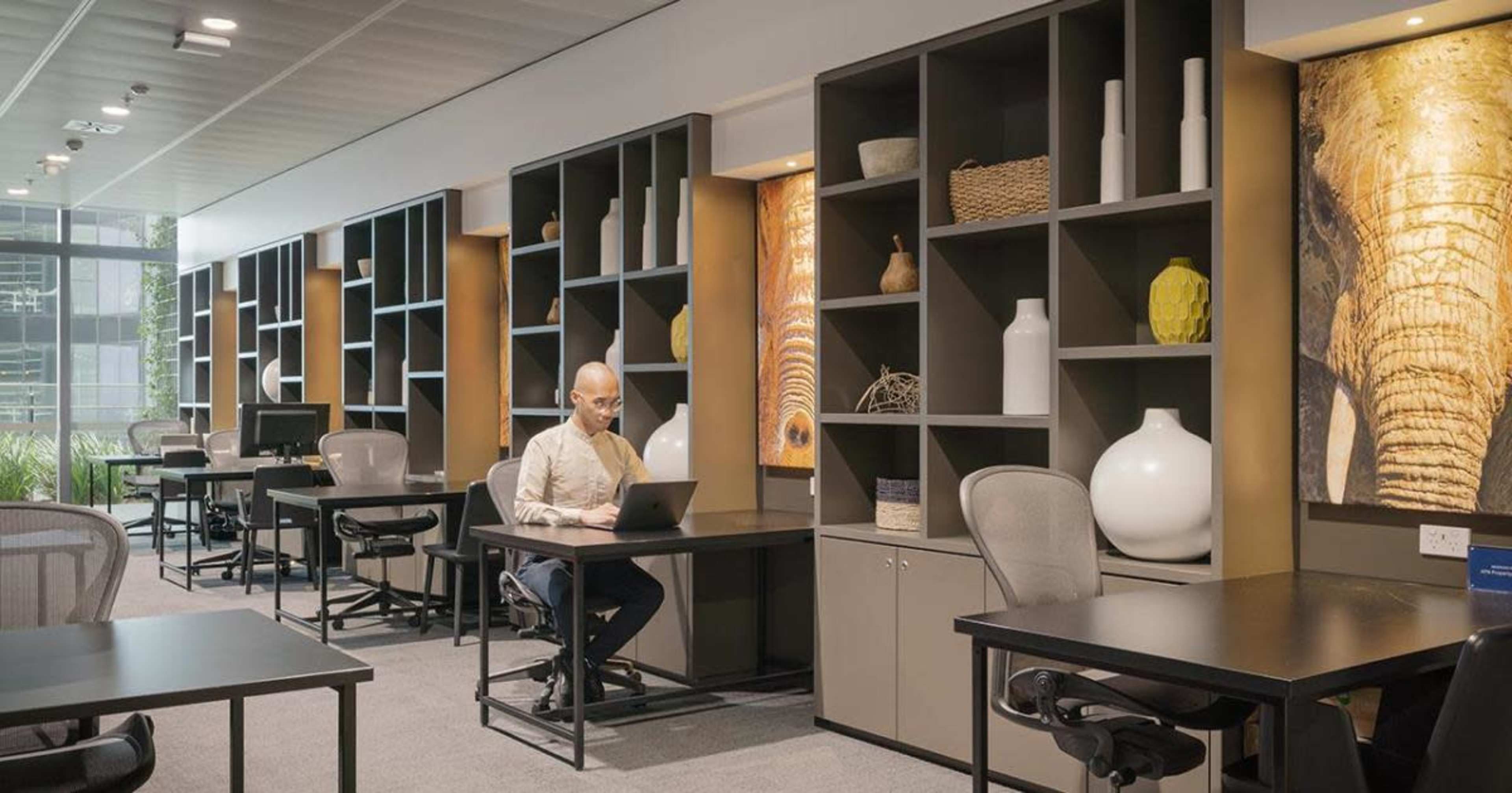A workspace features a man working on a laptop at a desk, surrounded by shelves displaying various decorative items.