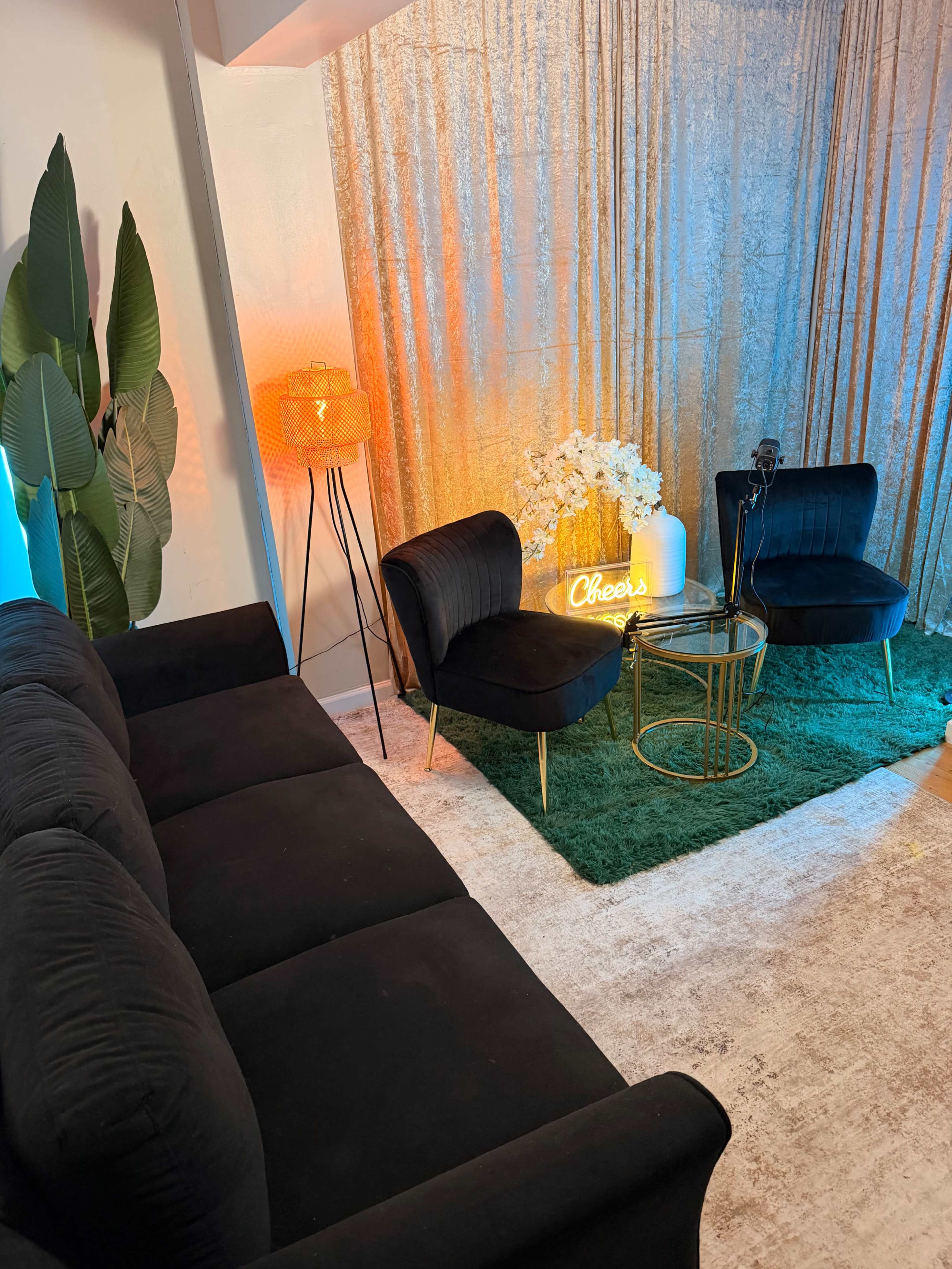 The image shows a cozy living area with a black sofa, two stylish chairs, a small circular table, and decorative elements including a lamp and a neon sign, all set against a backdrop of shimmering fabric.