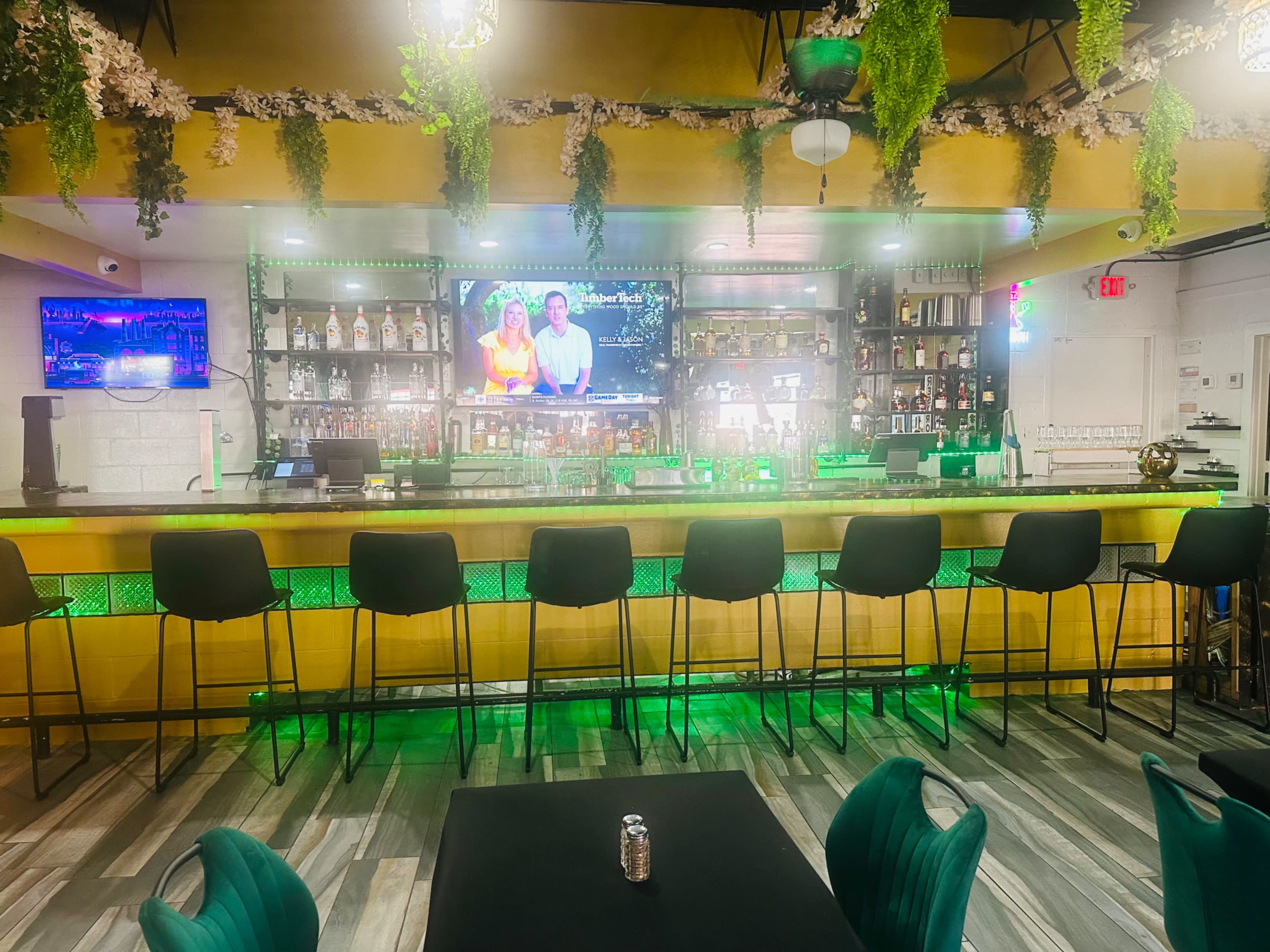 The image shows a modern bar area with a long counter, high stools, and a TV screen displaying a show, surrounded by decorative plants and ambient lighting.