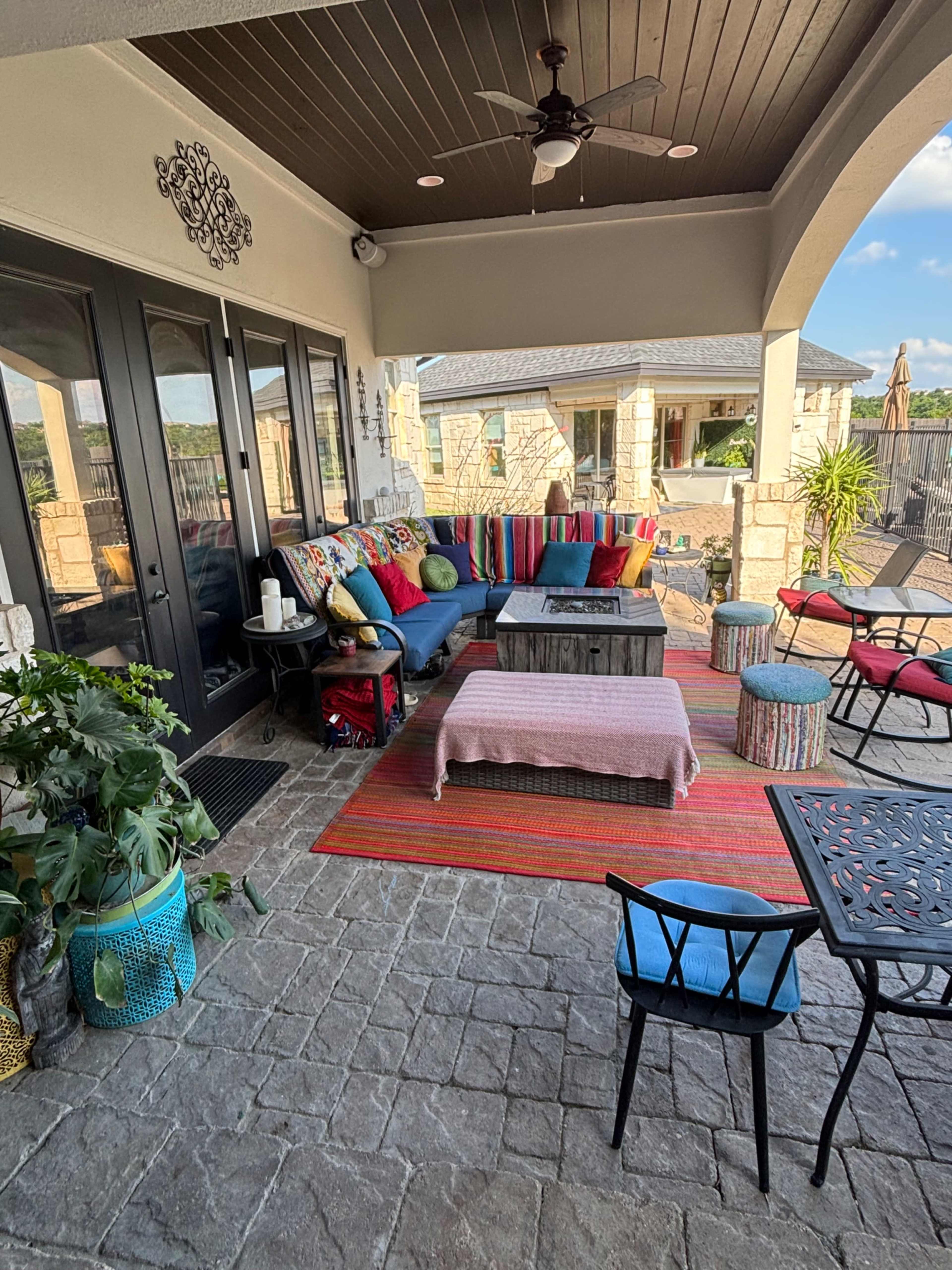 The image shows a stylish outdoor patio area furnished with colorful cushions, a wooden coffee table, and various seating options, surrounded by greenery and a view of the landscape.