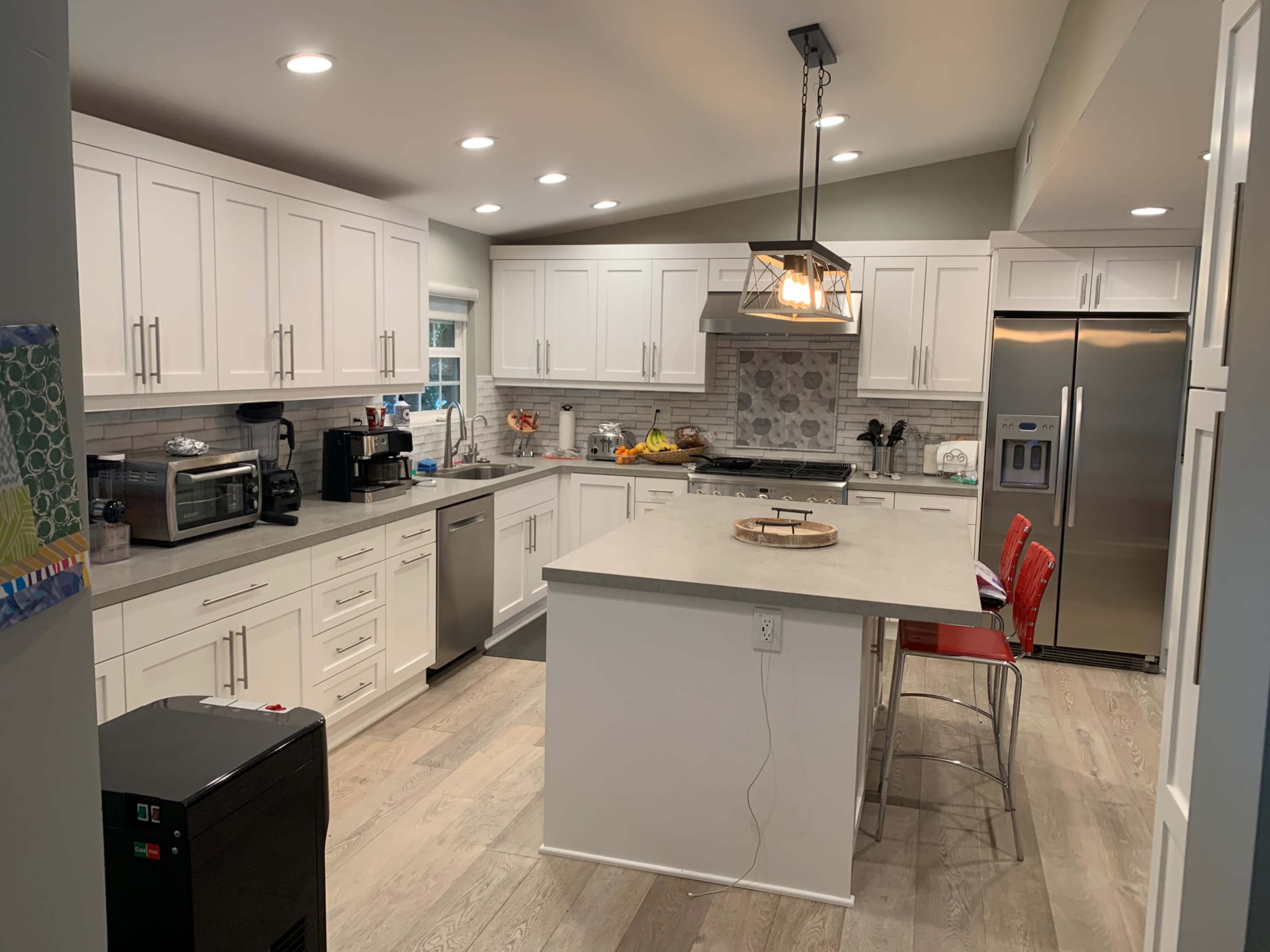 The image shows a modern kitchen with white cabinets, stainless steel appliances, and a central island with red bar stools.