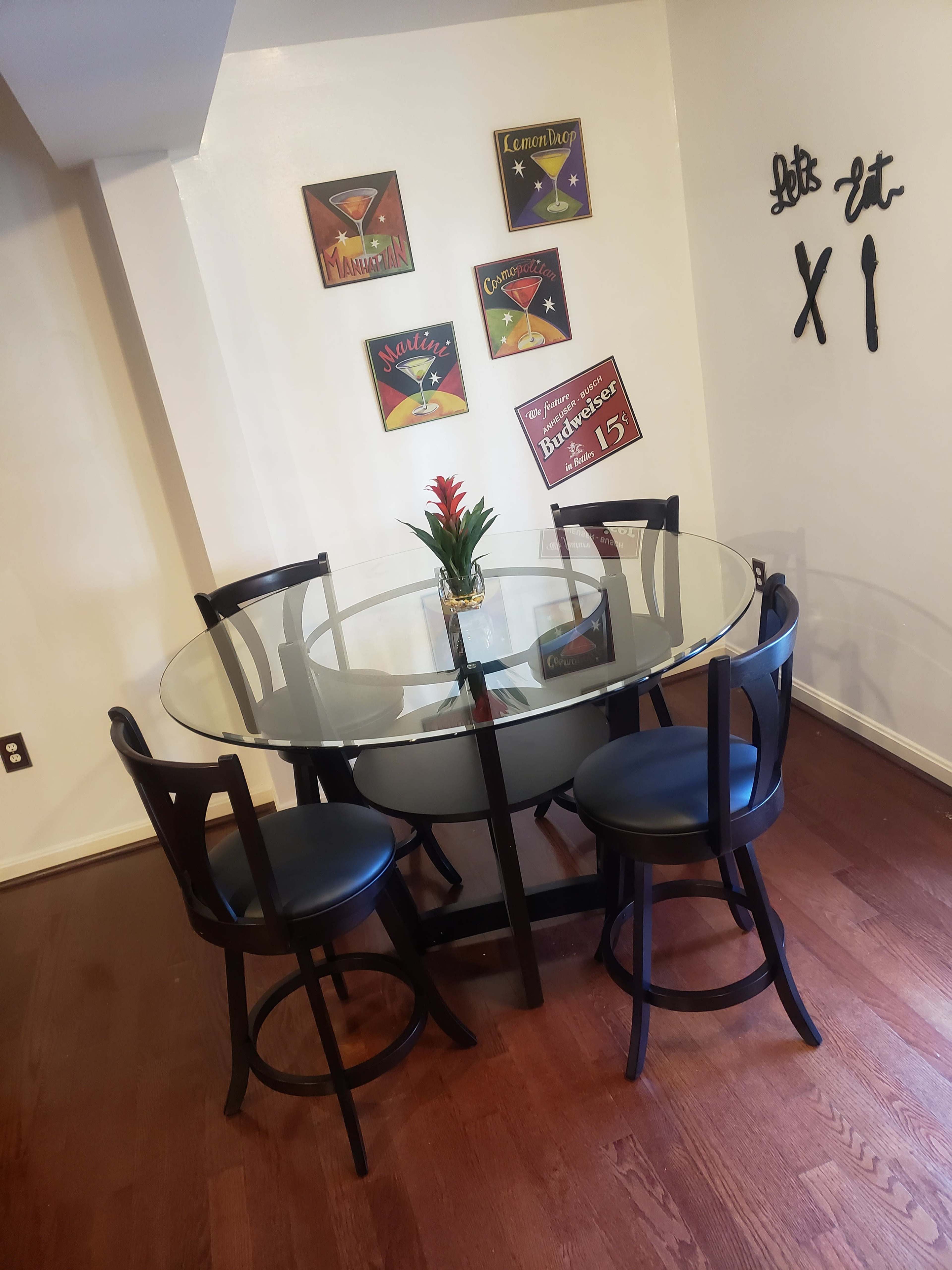 A glass-top dining table with four black chairs is set in a room adorned with colorful wall art and decor.