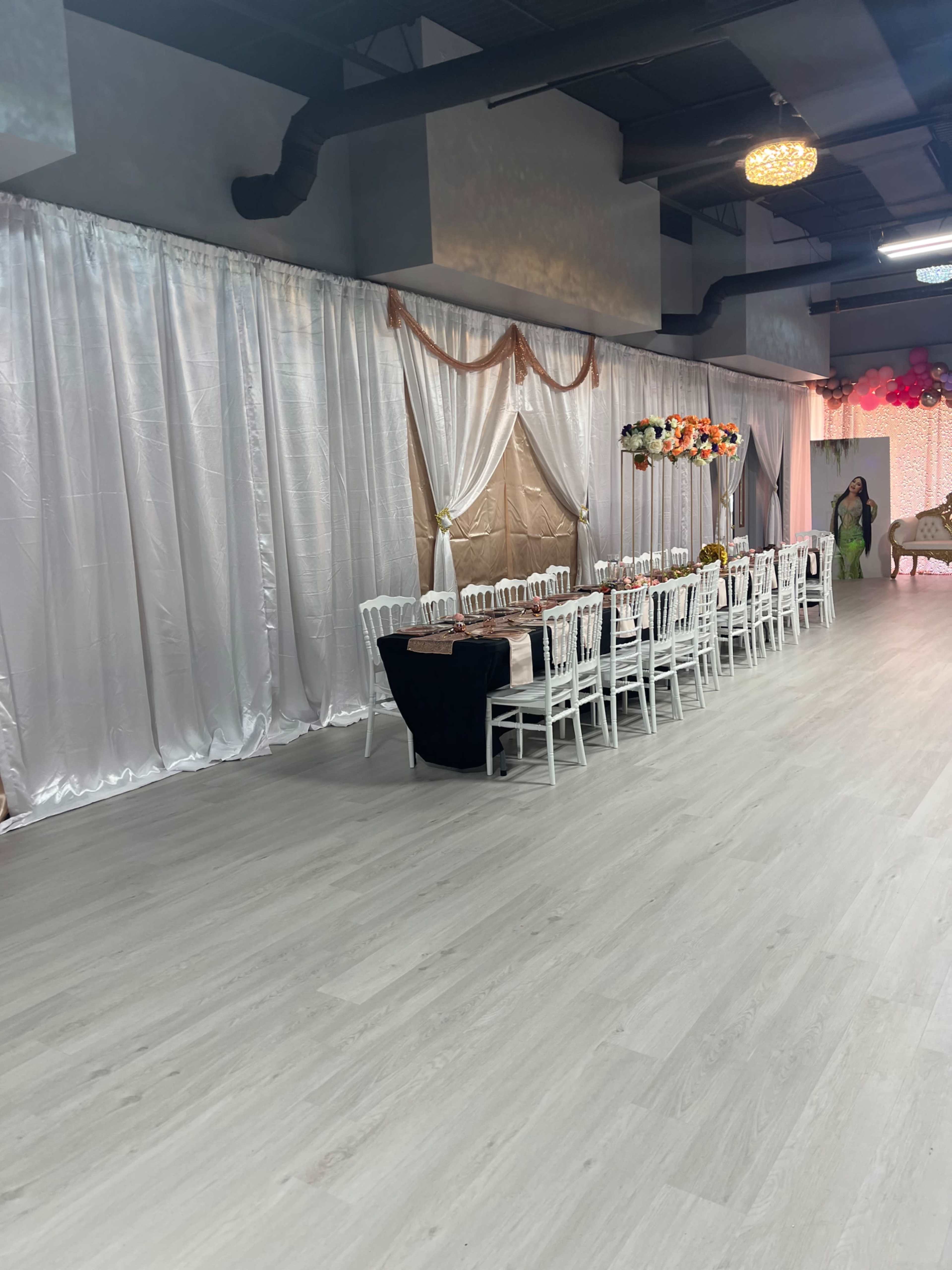 A spacious event venue with white draped curtains, a long banquet-style table set with white chairs, and decorative elements at one end.