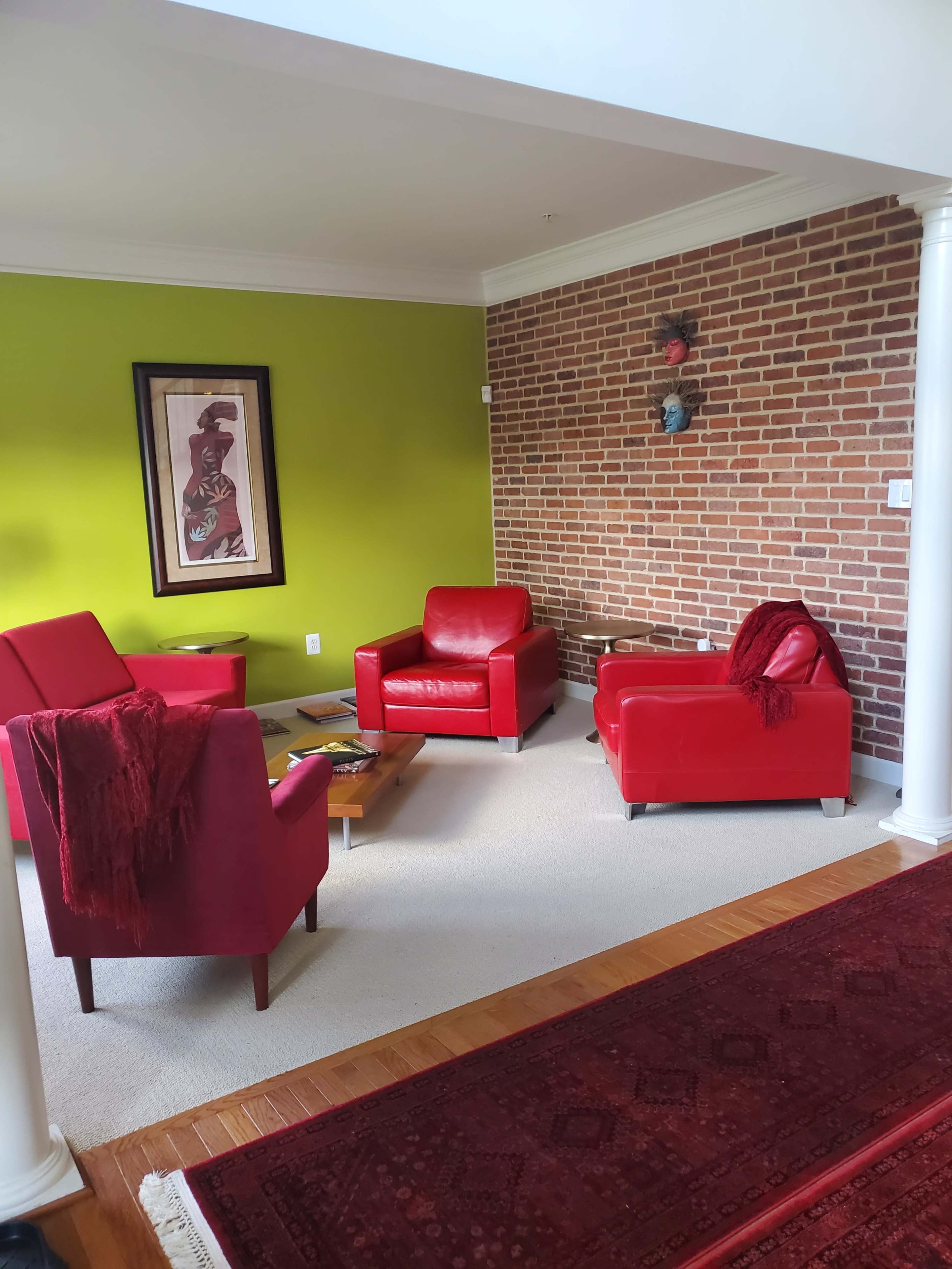 A bright living space with two red armchairs and a red ottoman, situated on a light carpet and next to a brick wall, with a green accent wall behind them.