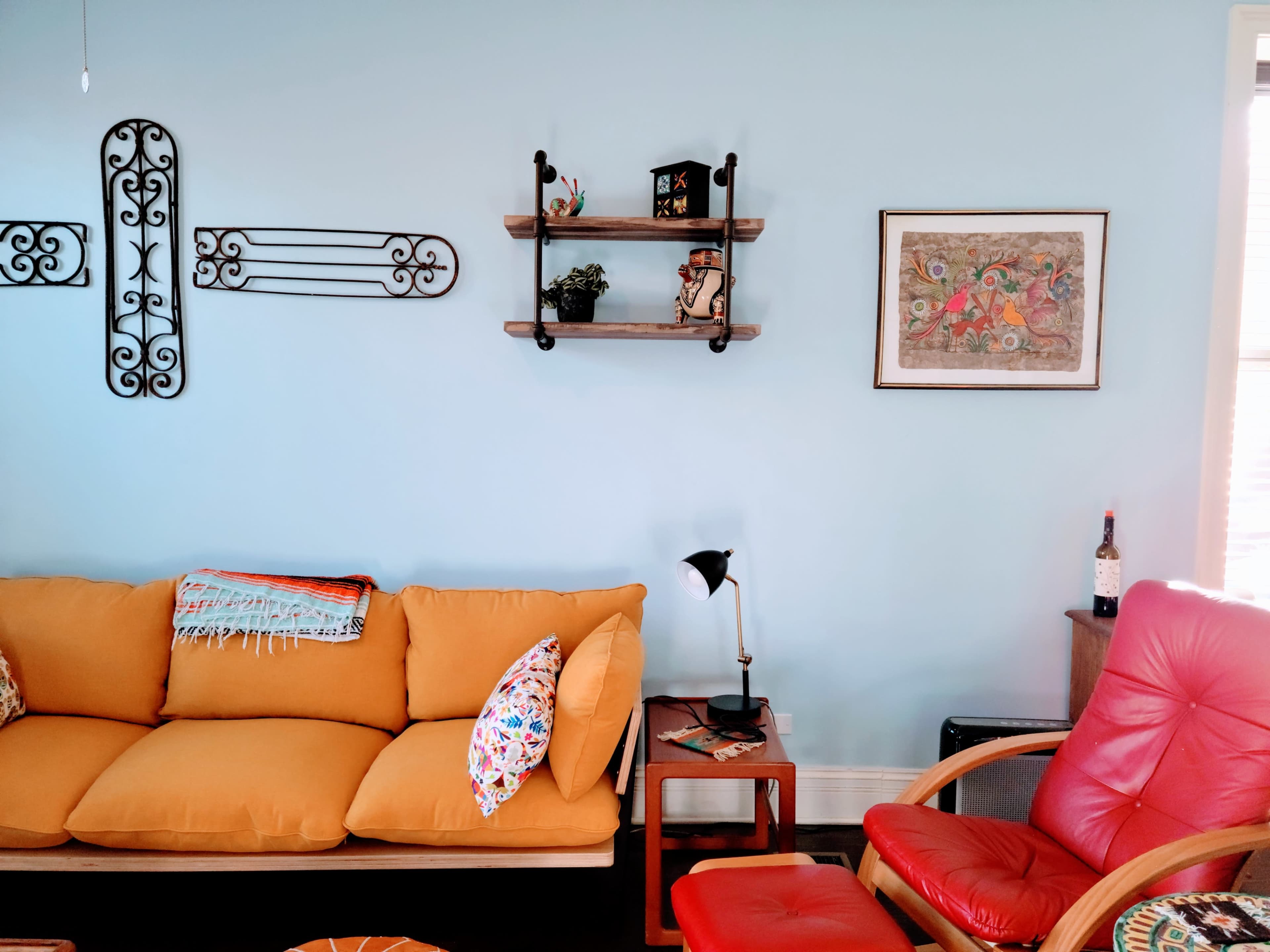 A living room with a bright blue wall, a yellow sofa, a red chair, shelves featuring decorative items, and a framed piece of art.
