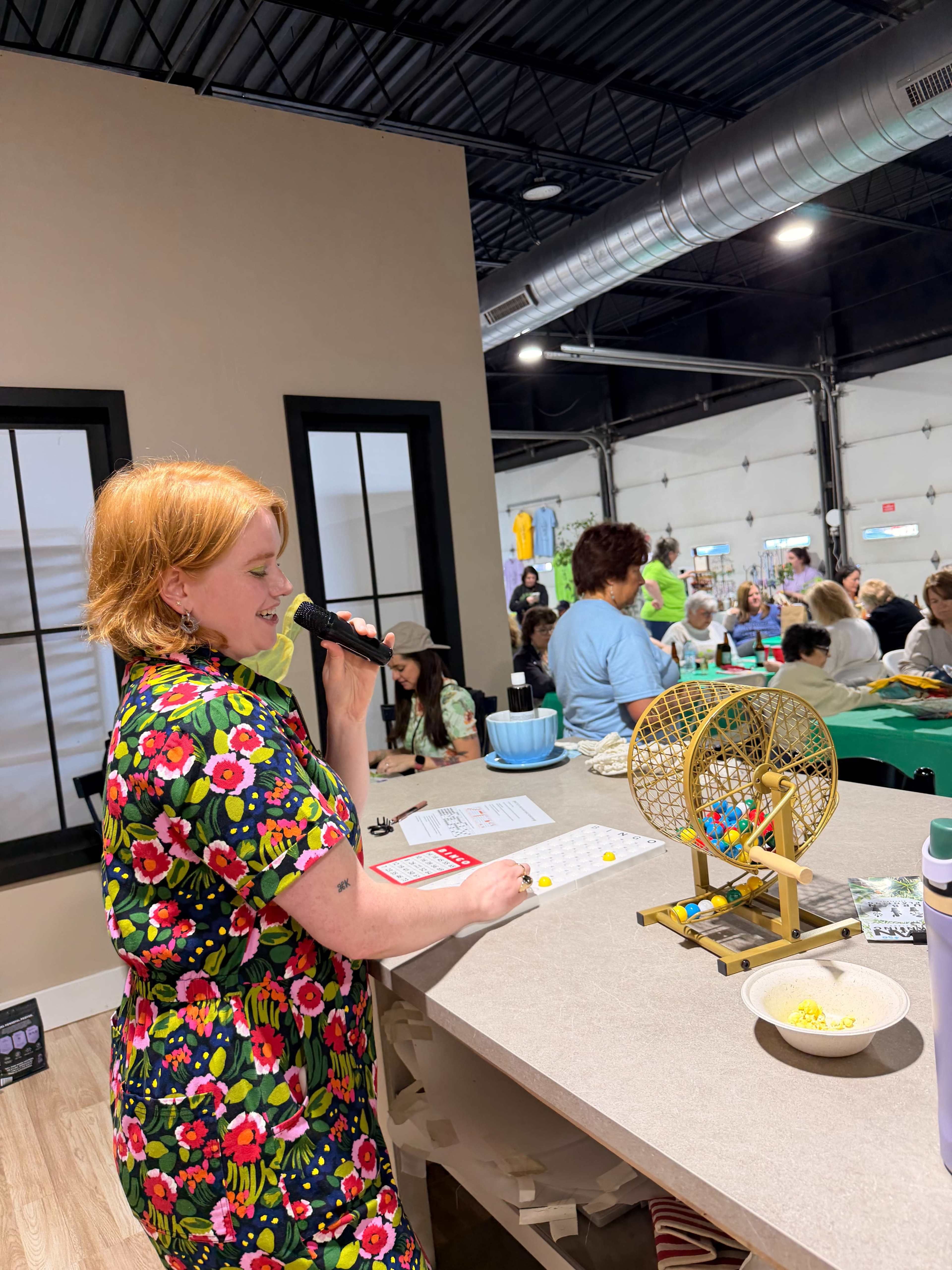 A woman with orange hair stands at a table, holding a microphone and overseeing a bingo game in a spacious community center filled with participants.