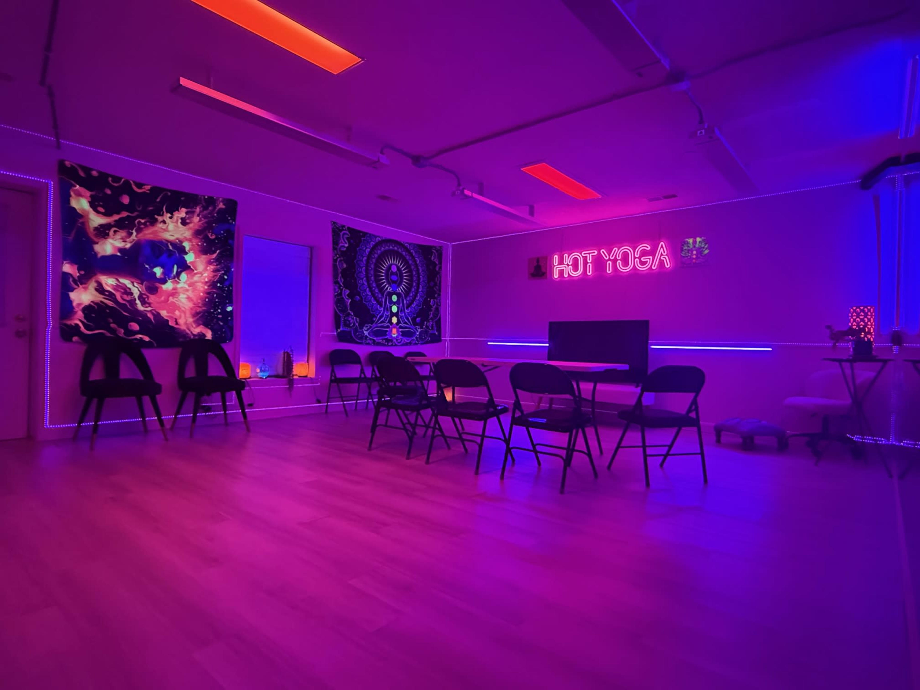 A brightly lit yoga studio features neon lighting, colorful wall art, and several folding chairs arranged around multiple tables.