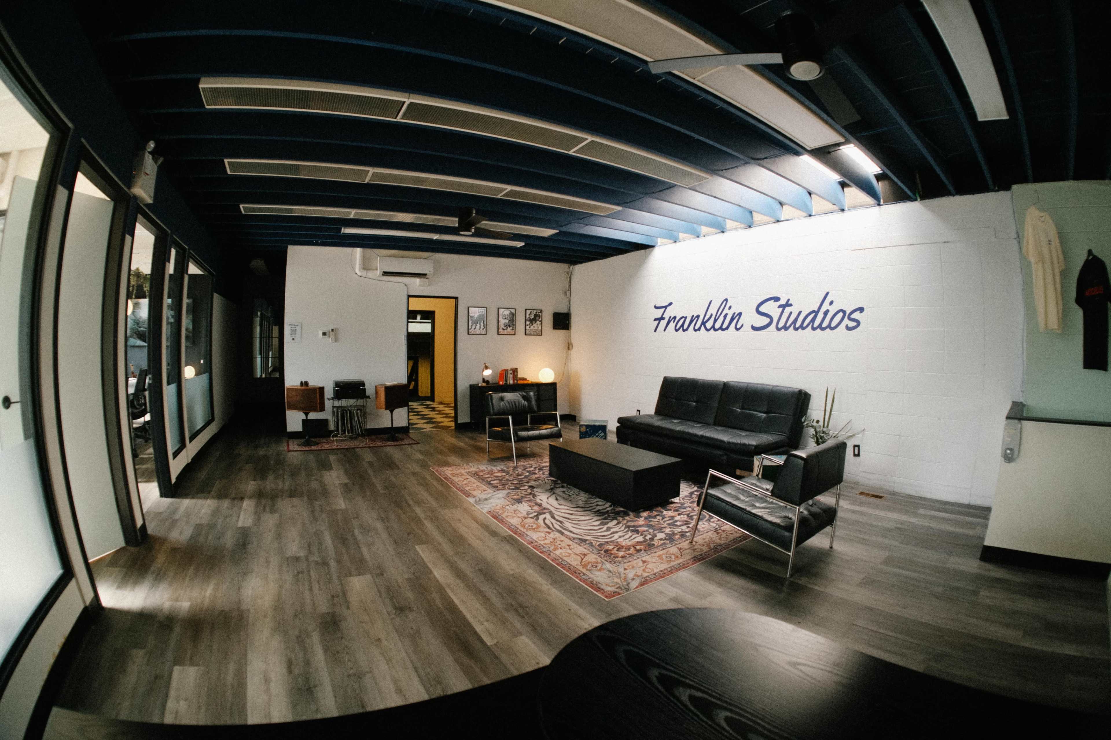 The image shows a modern office space featuring a black leather sofa, a coffee table, a patterned rug, and blue ceiling beams.