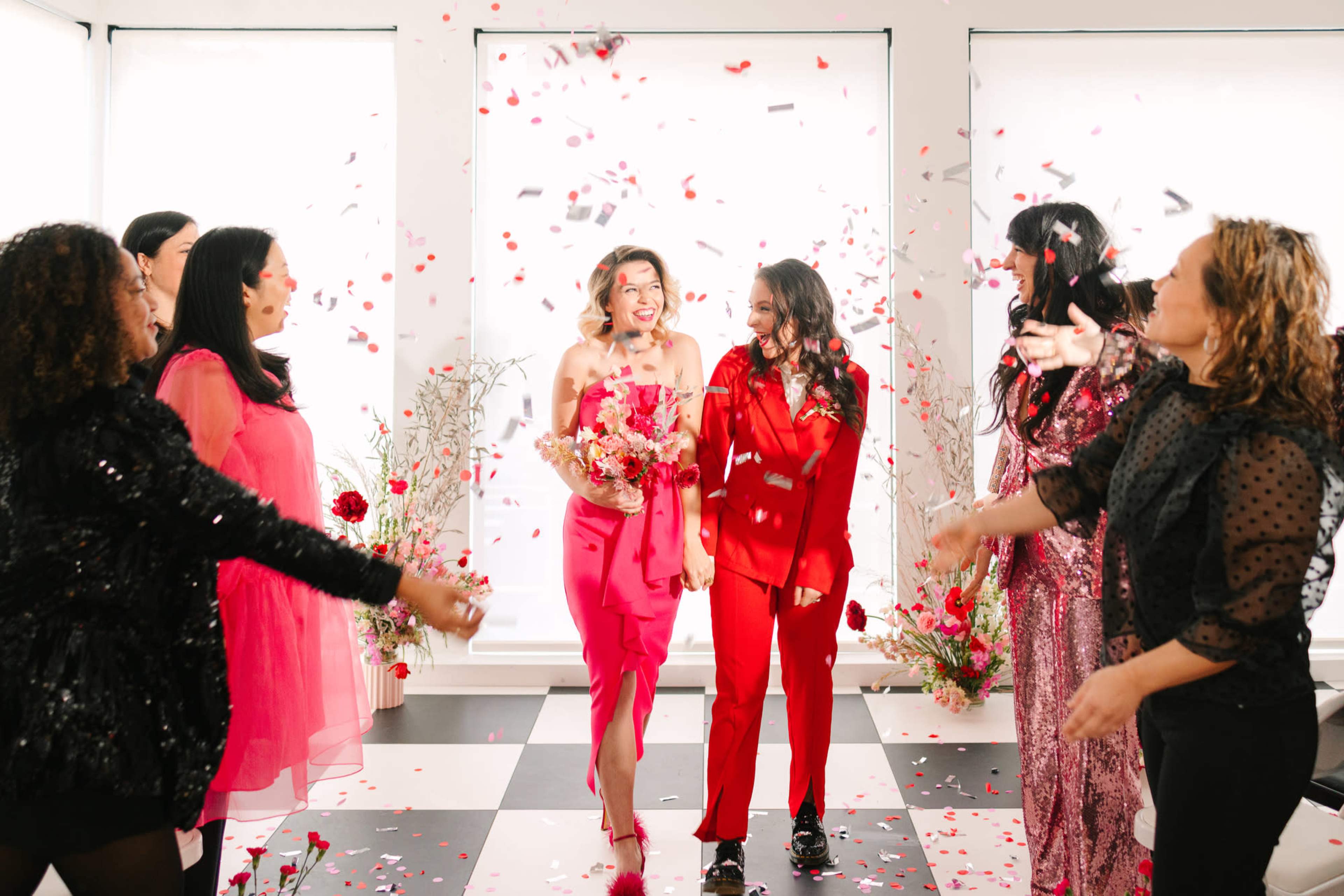 Two women, one in a pink dress and the other in a red outfit, stand in the center of a room as confetti and flower petals fall around them, surrounded by a group of smiling women.