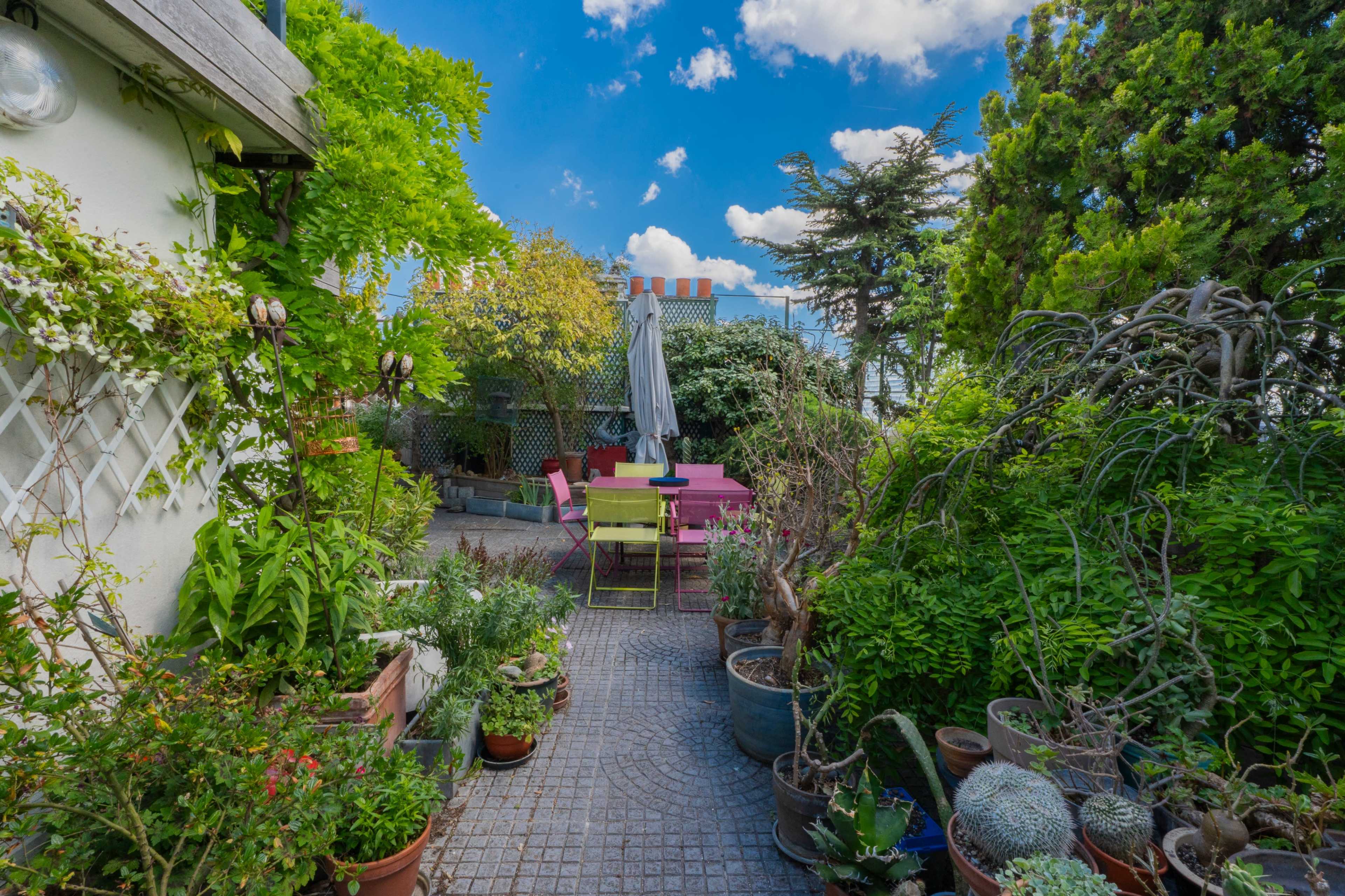 A vibrant garden space features potted plants and seating areas under a clear blue sky with scattered clouds.
