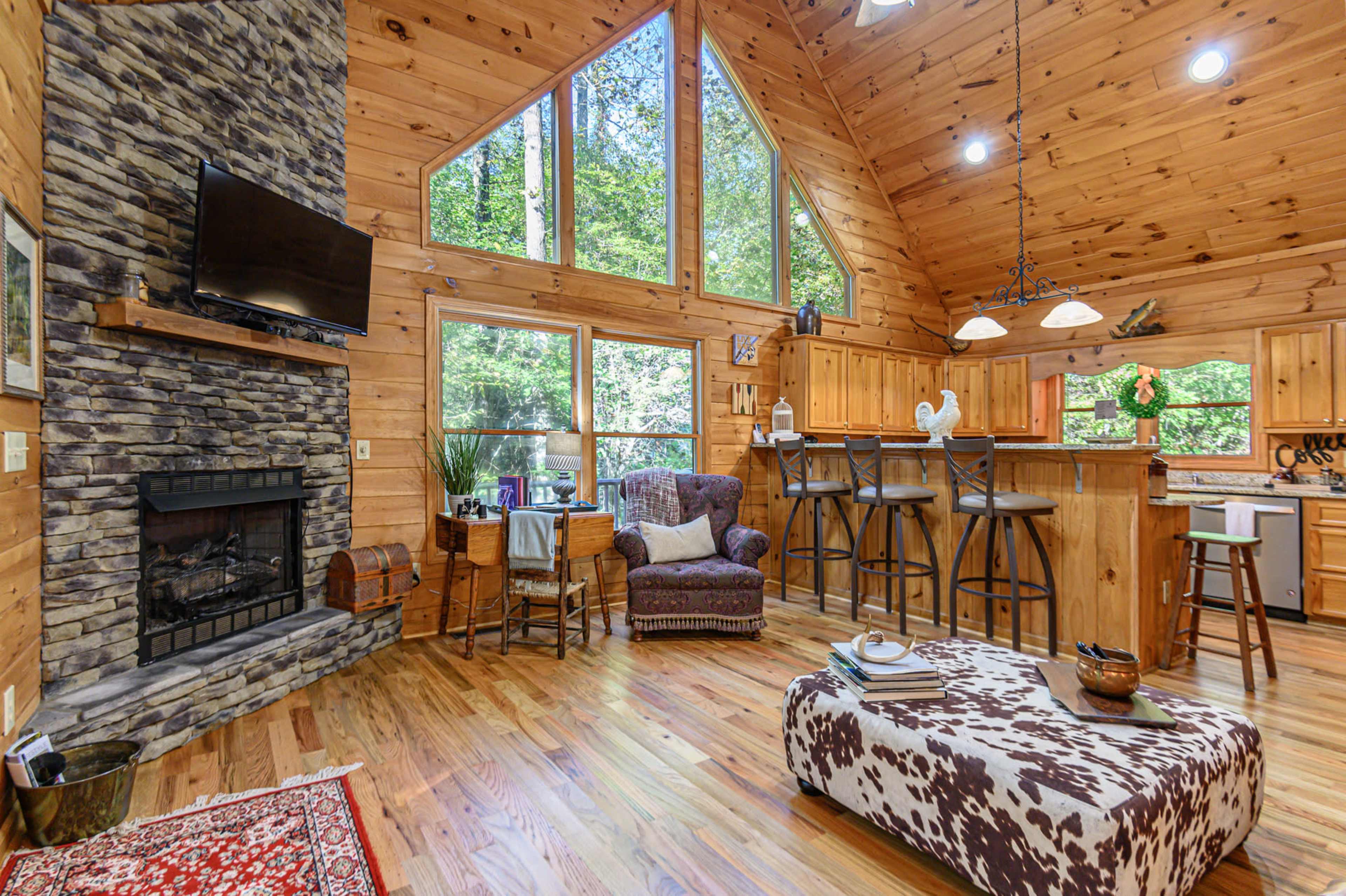 The spacious living area features large windows, a stone fireplace, wooden walls, and a mix of seating and dining arrangements.