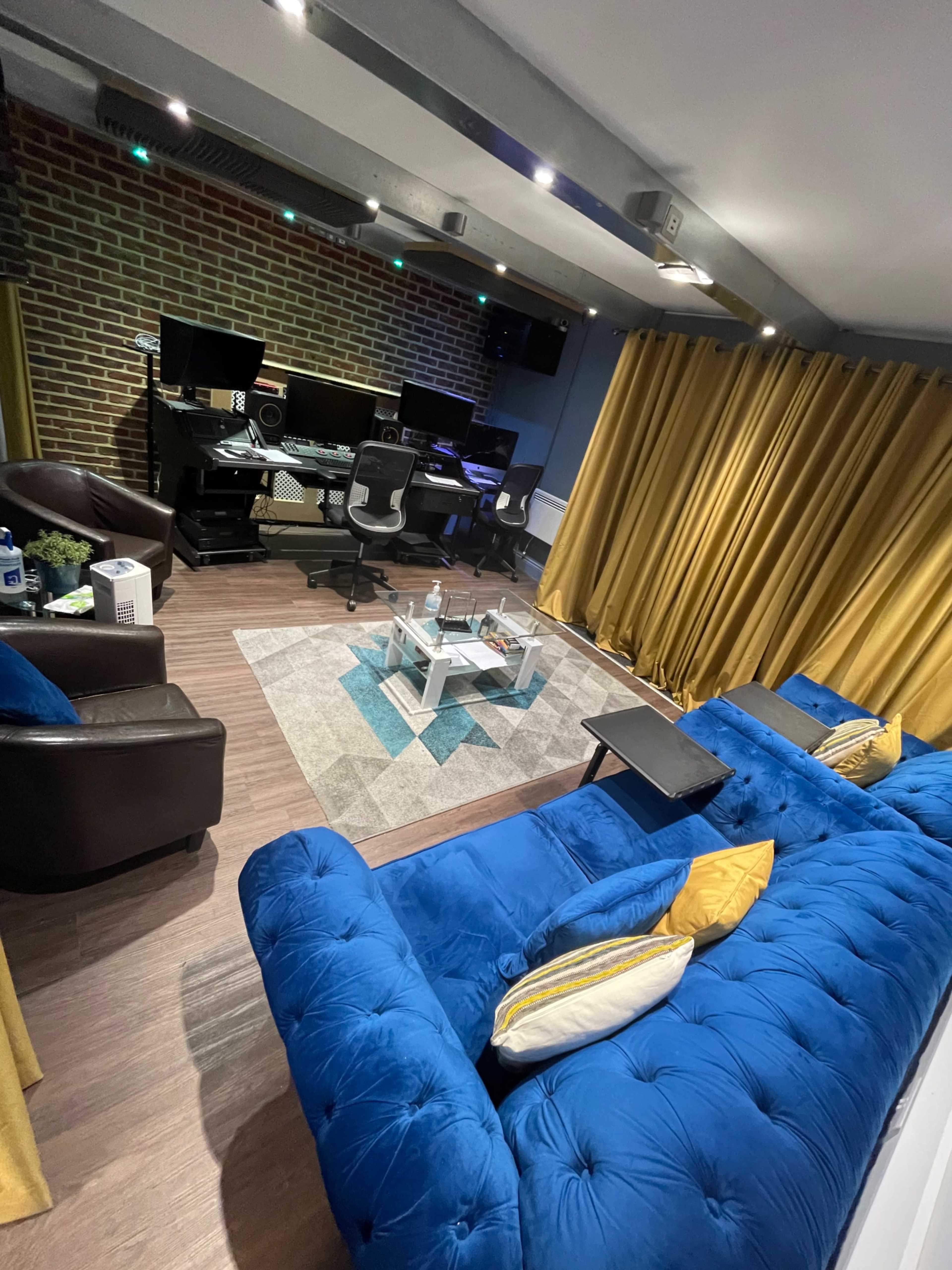 A modern recording studio featuring a blue velvet sectional sofa, a glass coffee table, and a workstation with multiple monitors against a brick wall, complemented by yellow curtains.