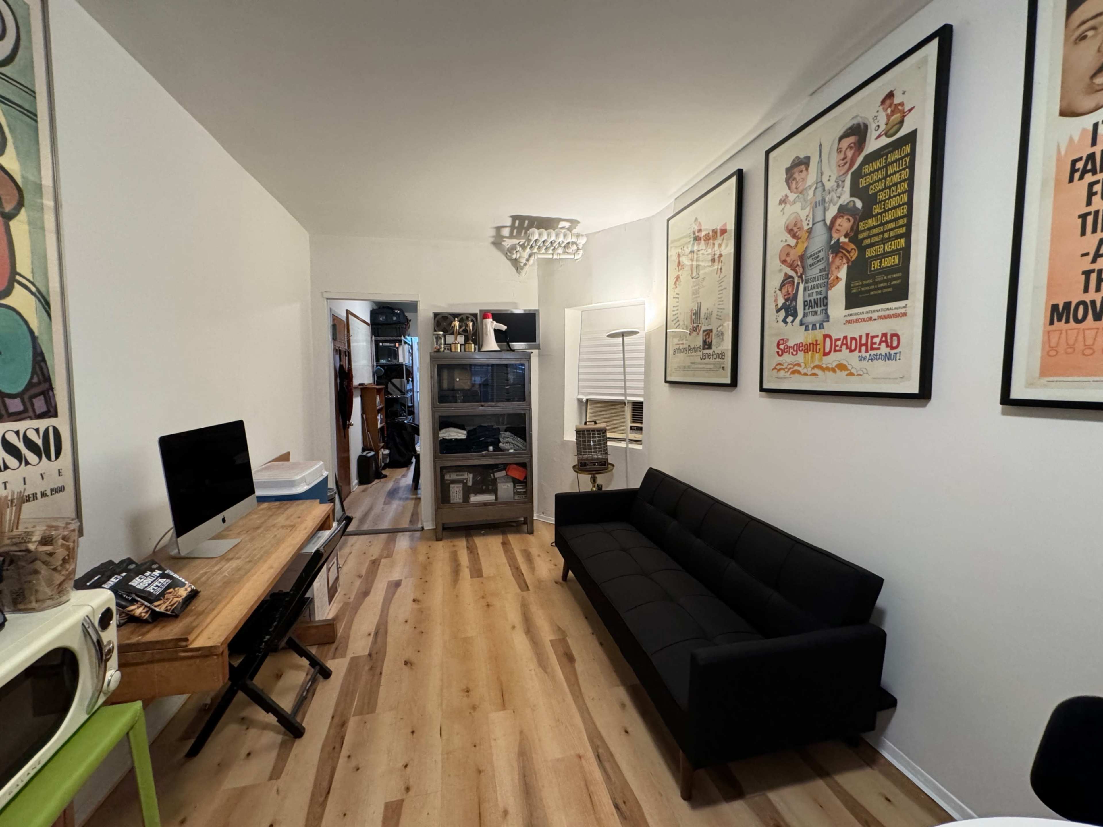 The image shows a living room featuring a black sofa, a wooden table, and several framed posters on the walls, with a doorway leading to another room.