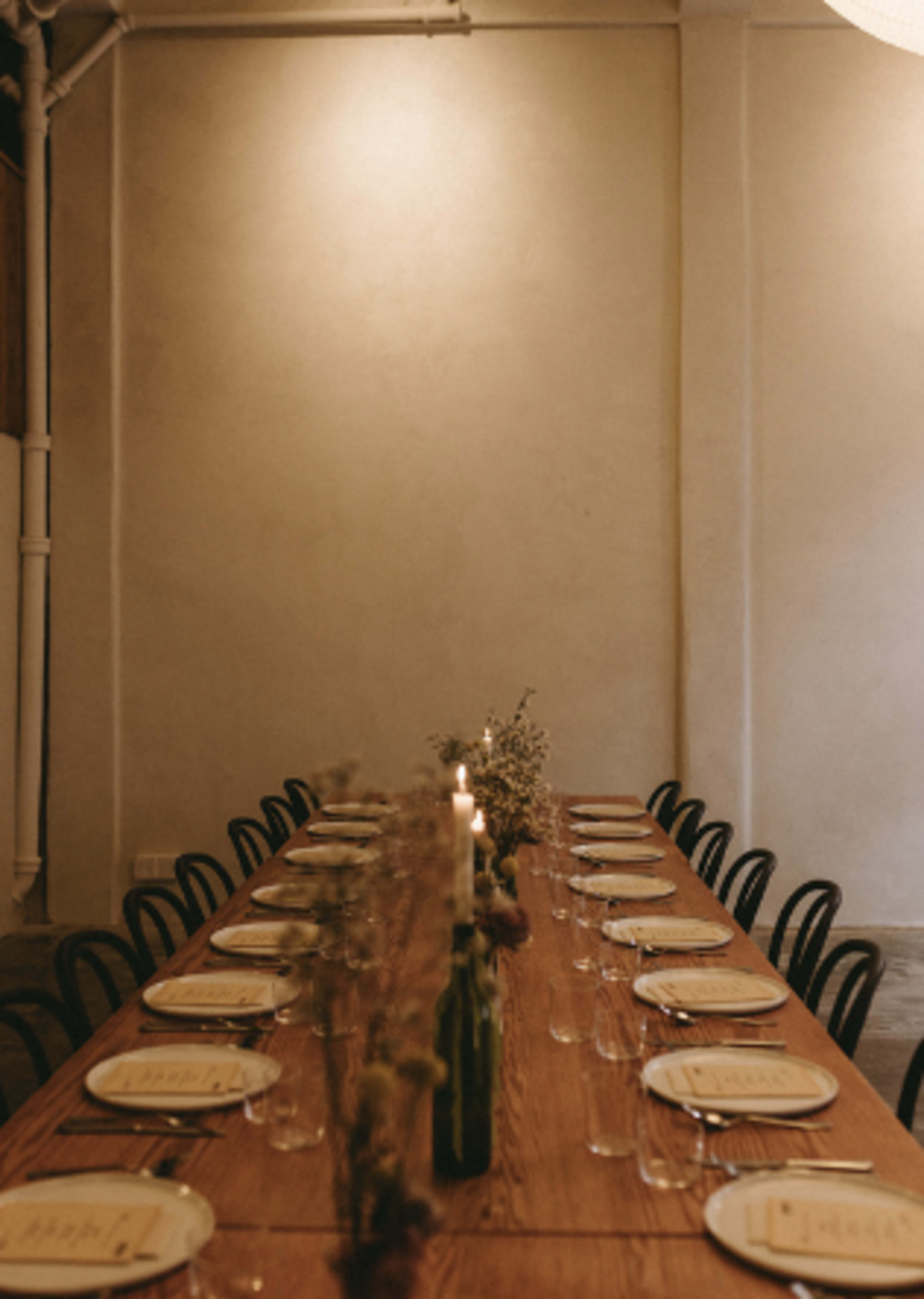A long wooden dining table is set with plates and glasses, featuring a simple floral centerpiece and candlelight along the center.