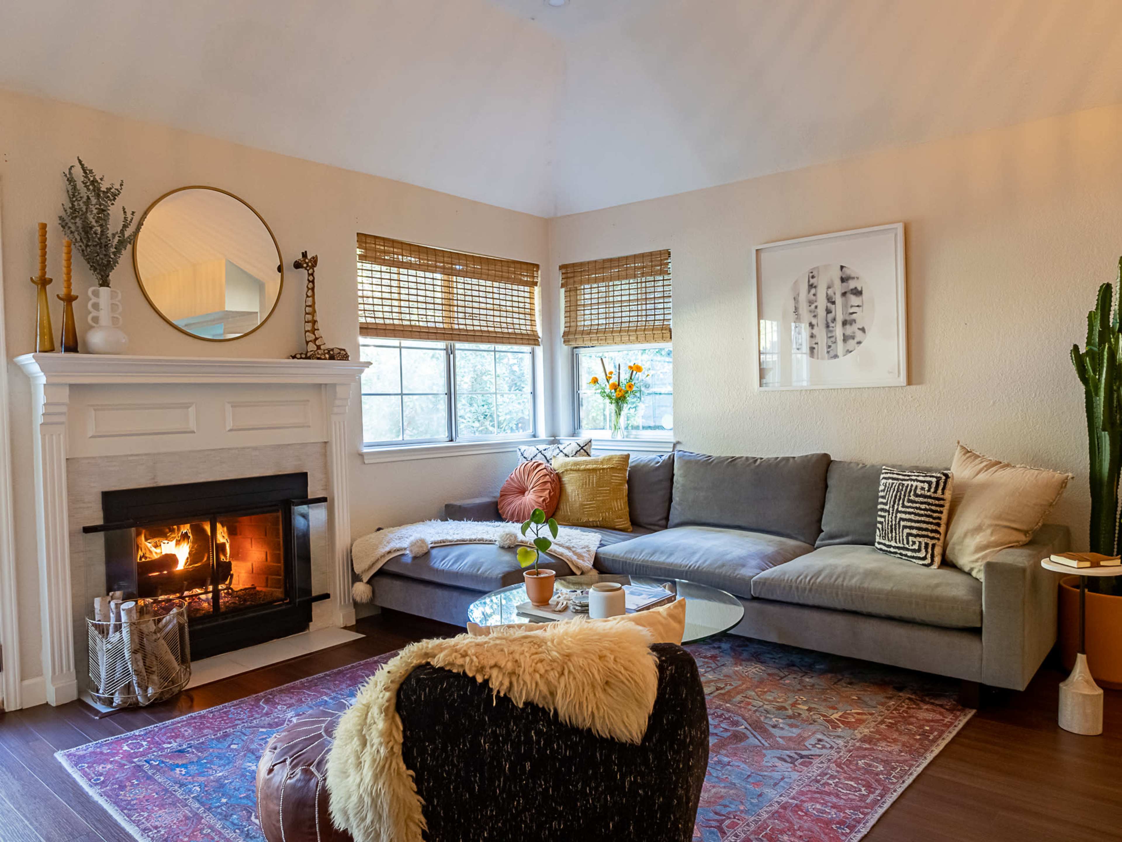 A cozy living room with a sectional sofa, a fireplace with a fire burning, and decorative elements like a round mirror and potted plants.