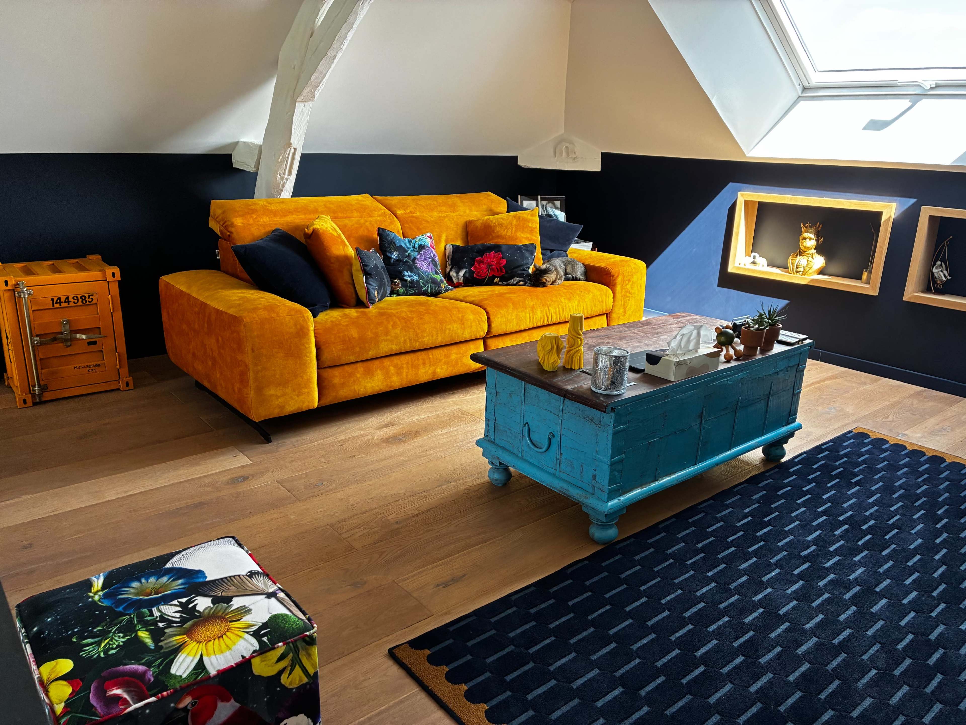 The image shows a cozy living room with an orange sofa, a blue coffee table, and decorative shelves against a partially dark blue wall.