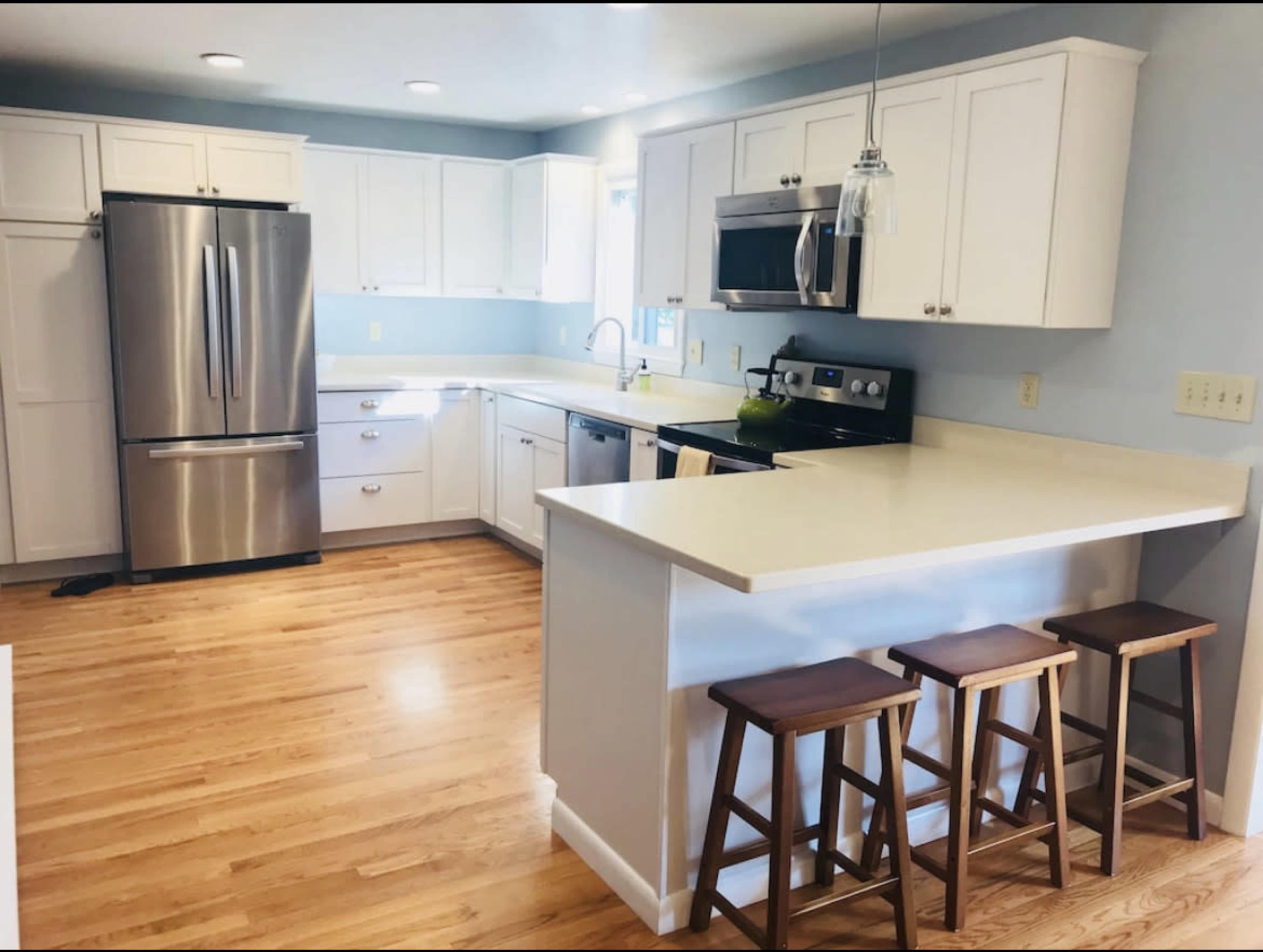 A modern kitchen features stainless steel appliances, white cabinetry, a light-colored countertop, and three wooden stools at a breakfast bar.