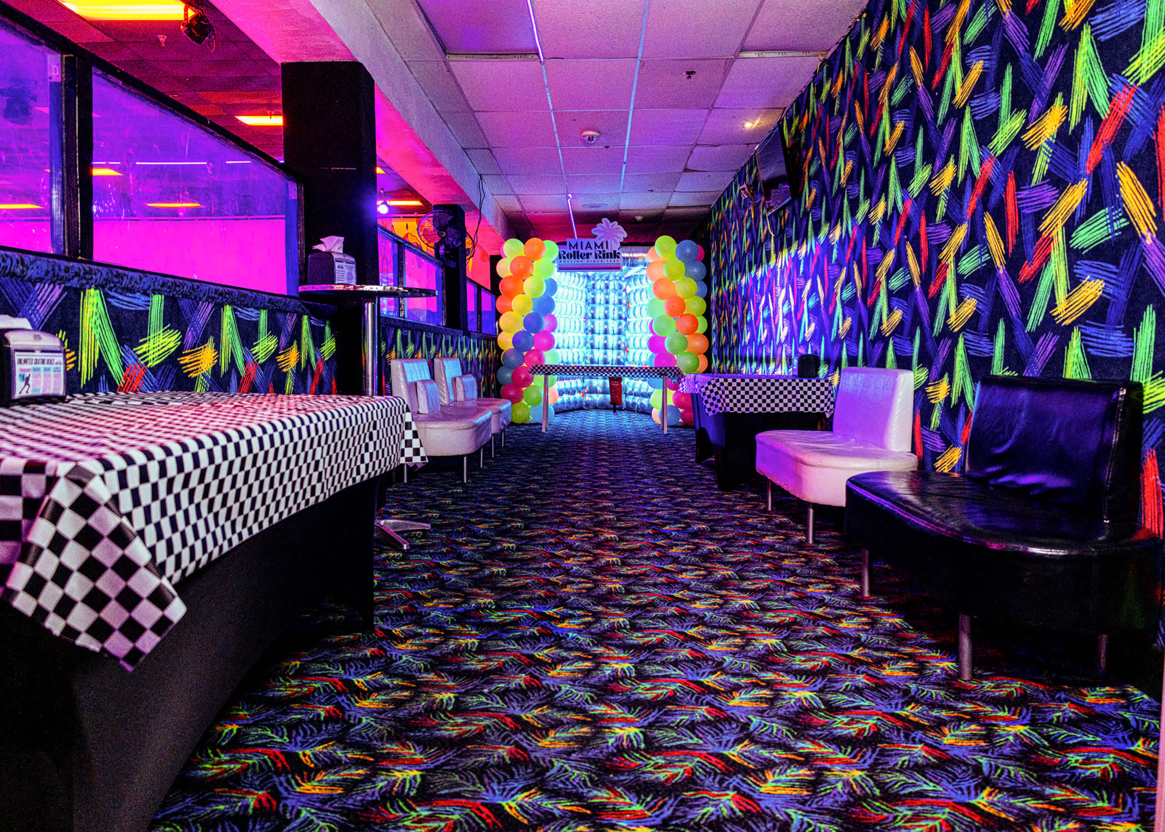 The image shows a brightly colored hallway with patterned walls, a checkered table, and a balloon archway at the end.