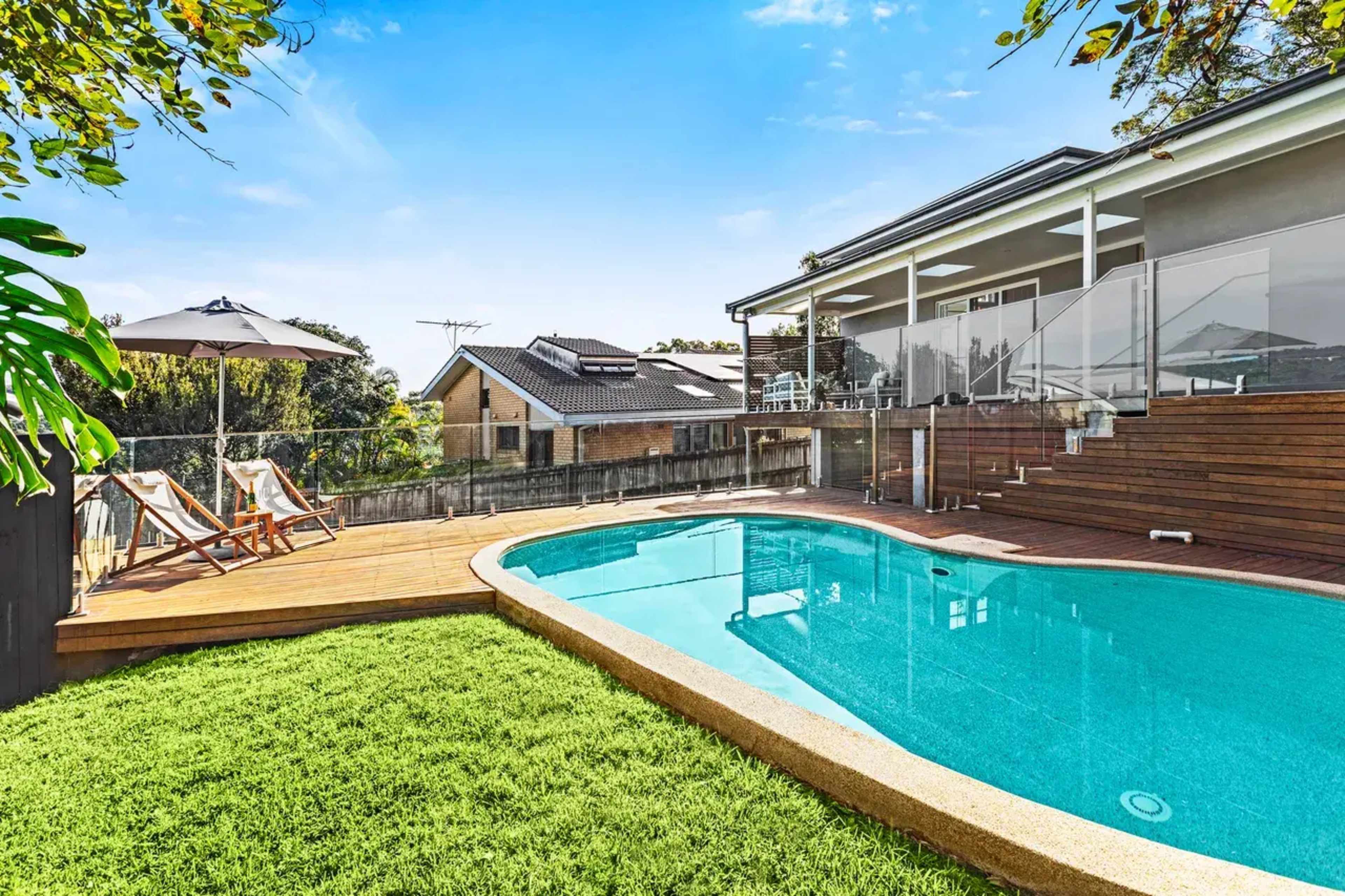 A residential backyard featuring a swimming pool surrounded by a wooden deck and lawn, with a house and outdoor furniture in the background.