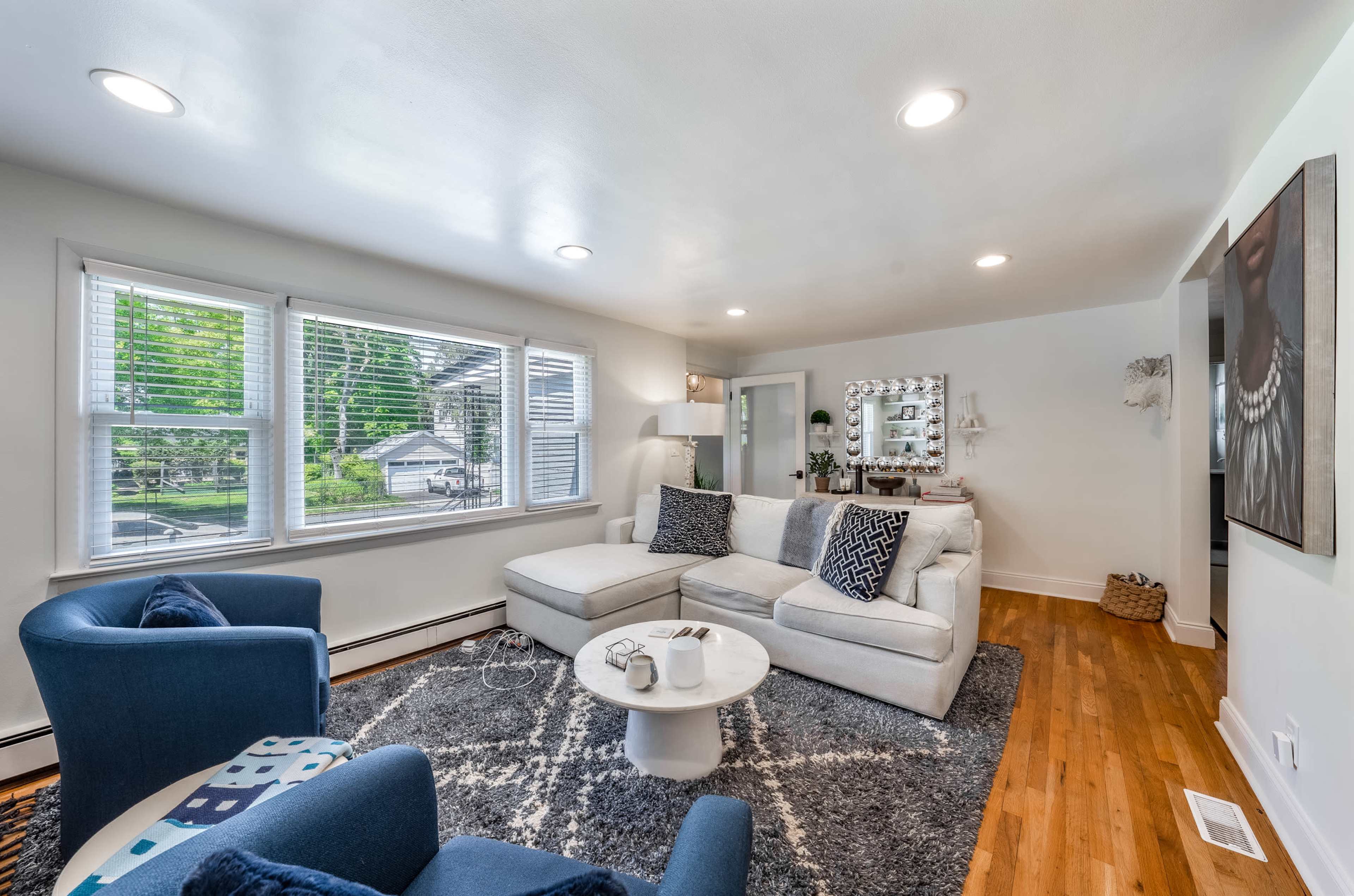 A modern living room featuring a light-colored sectional sofa, blue accent chairs, and a round area rug, with large windows allowing natural light to enter.