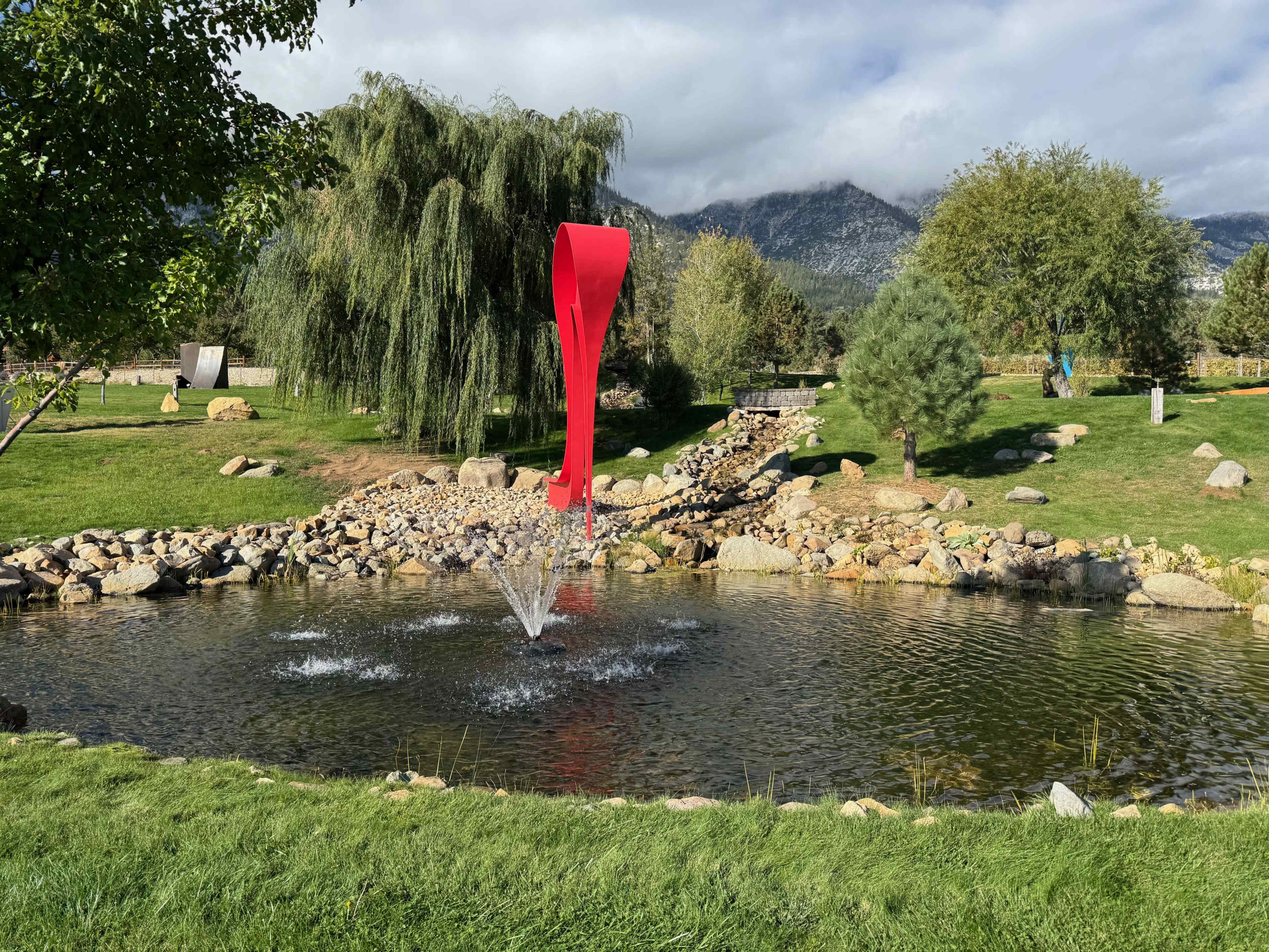 A red sculpture stands near a pond with a fountain, surrounded by trees and rocks against a backdrop of mountains and cloudy skies.