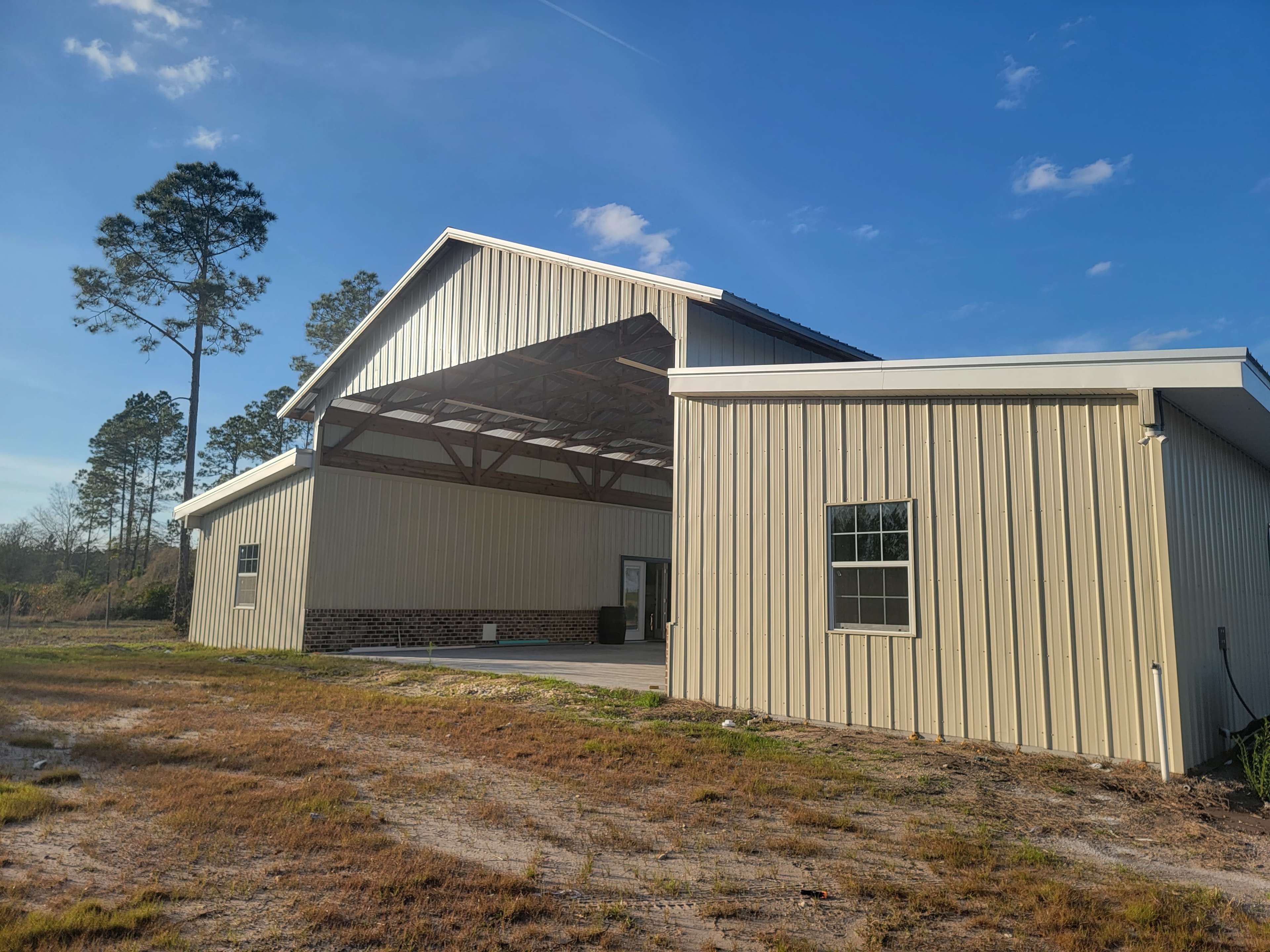 A large, open-sided metal building with a connected lower structure, surrounded by grass and trees under a clear blue sky.