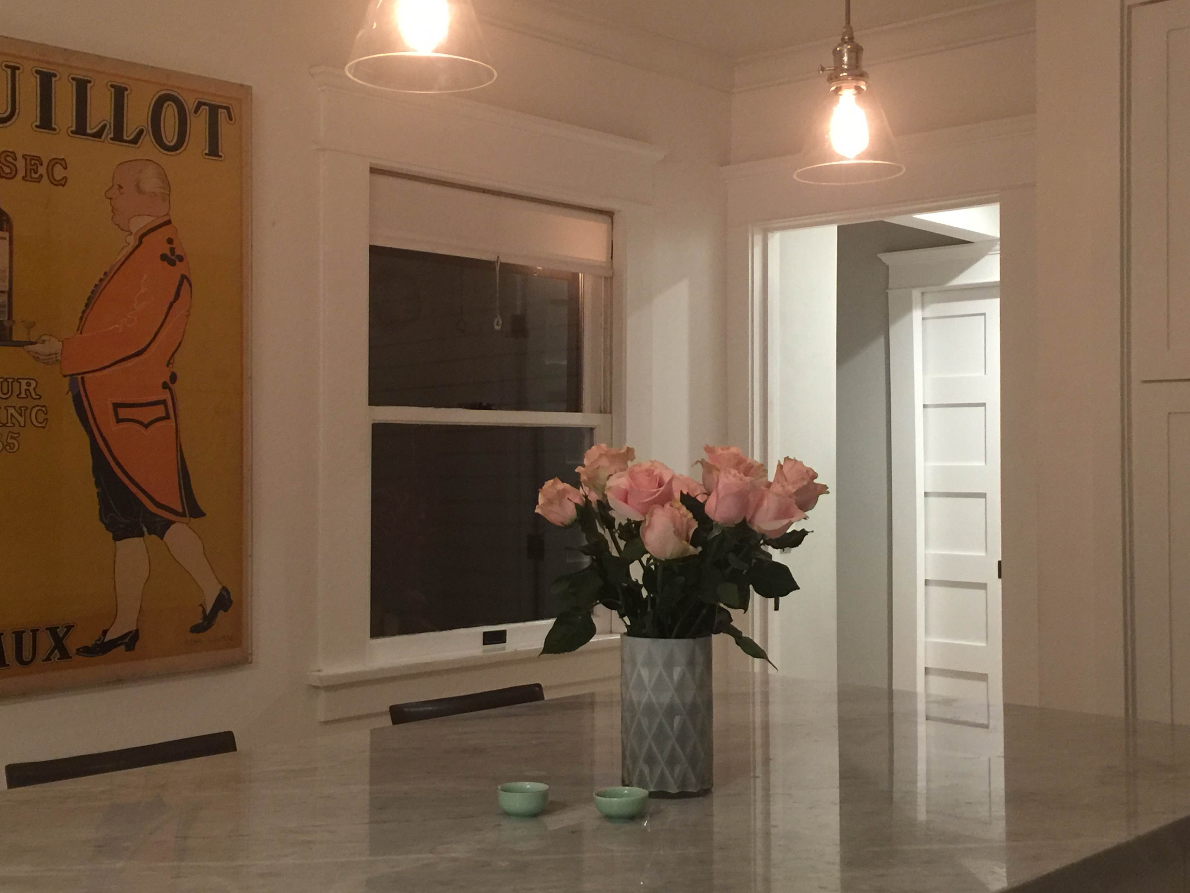 A marble kitchen countertop features a vase of pink roses, with a vintage poster and illuminated pendant lights in the background.