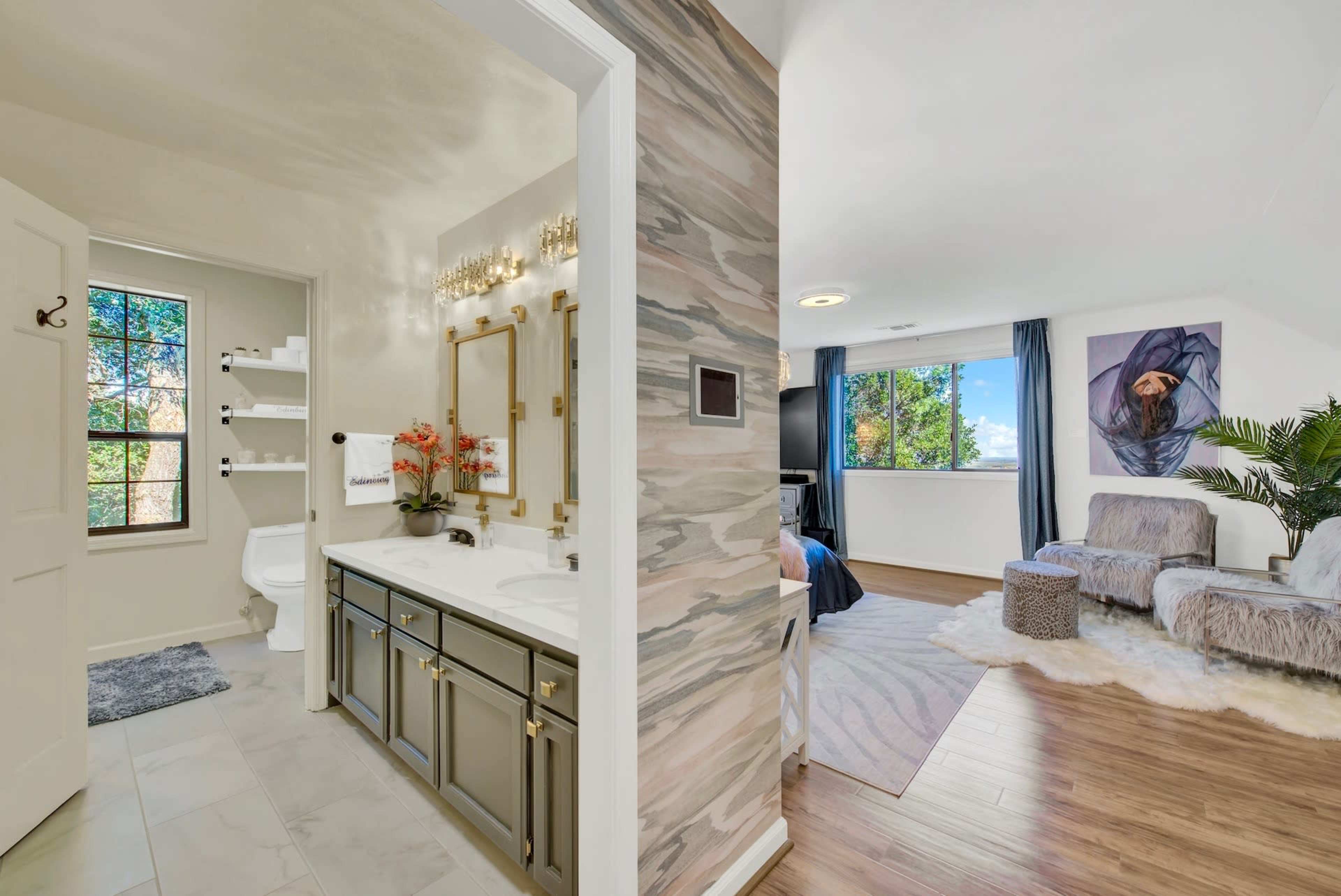 A well-lit bathroom connected to a bedroom, featuring a dual-sink vanity, a toilet, and a cozy seating area with window views.