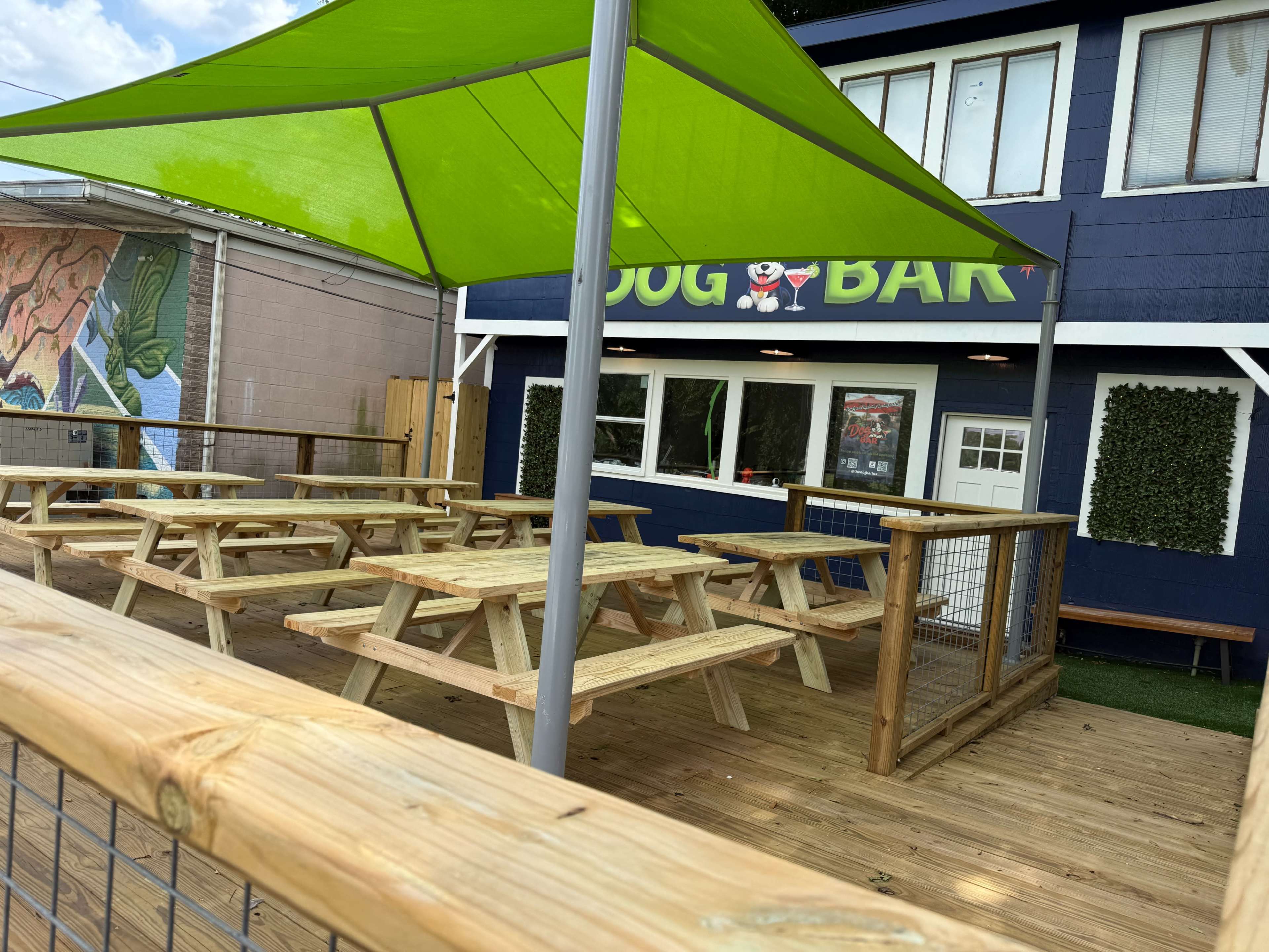 The image shows an outdoor seating area with wooden picnic tables beneath a green canopy, adjacent to a building labeled as a "Dog Bar."