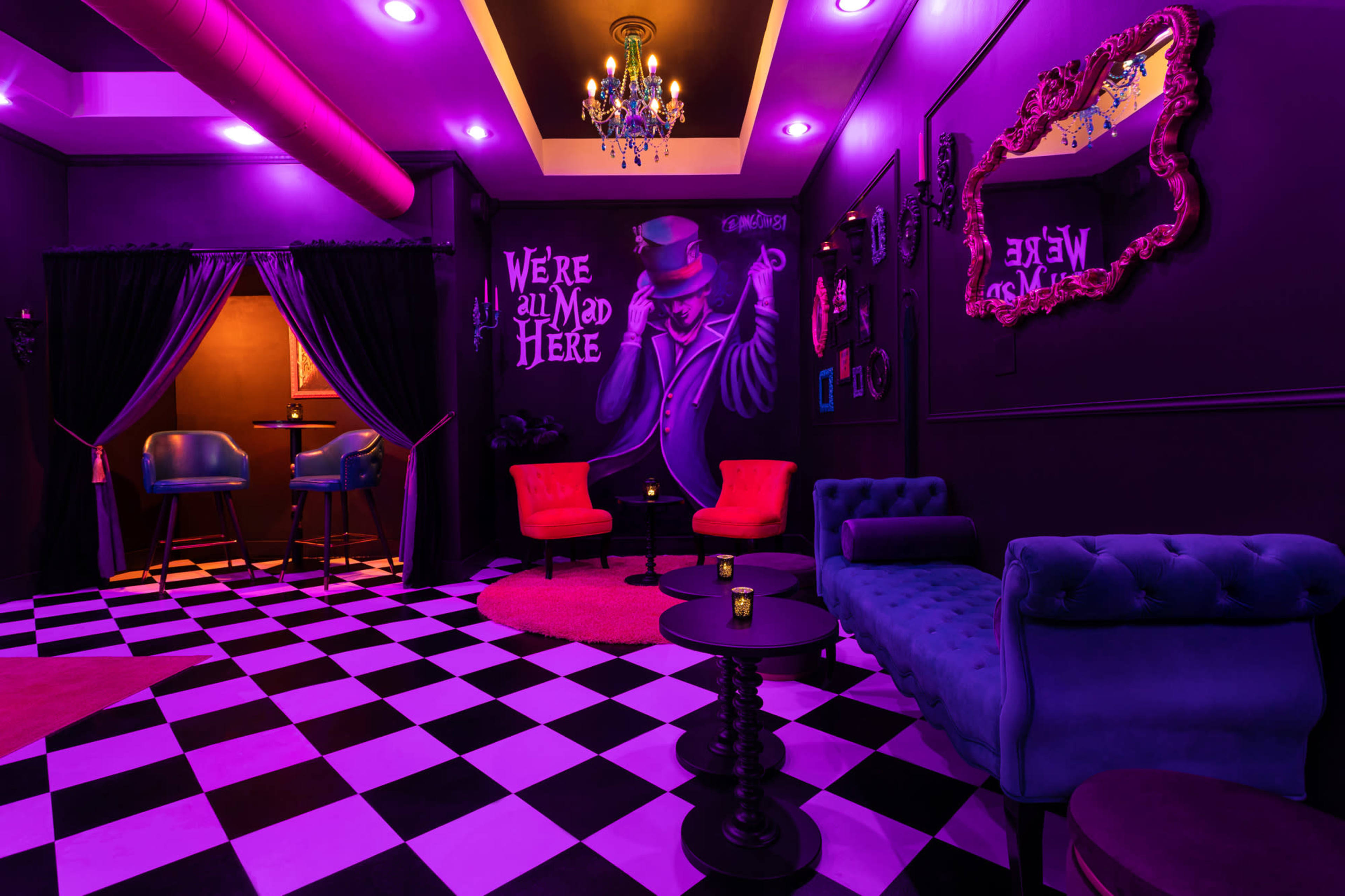 The image depicts a dimly lit lounge with a checkered floor, featuring purple walls, stylish furniture, and a whimsical mural that says "We're all mad here."